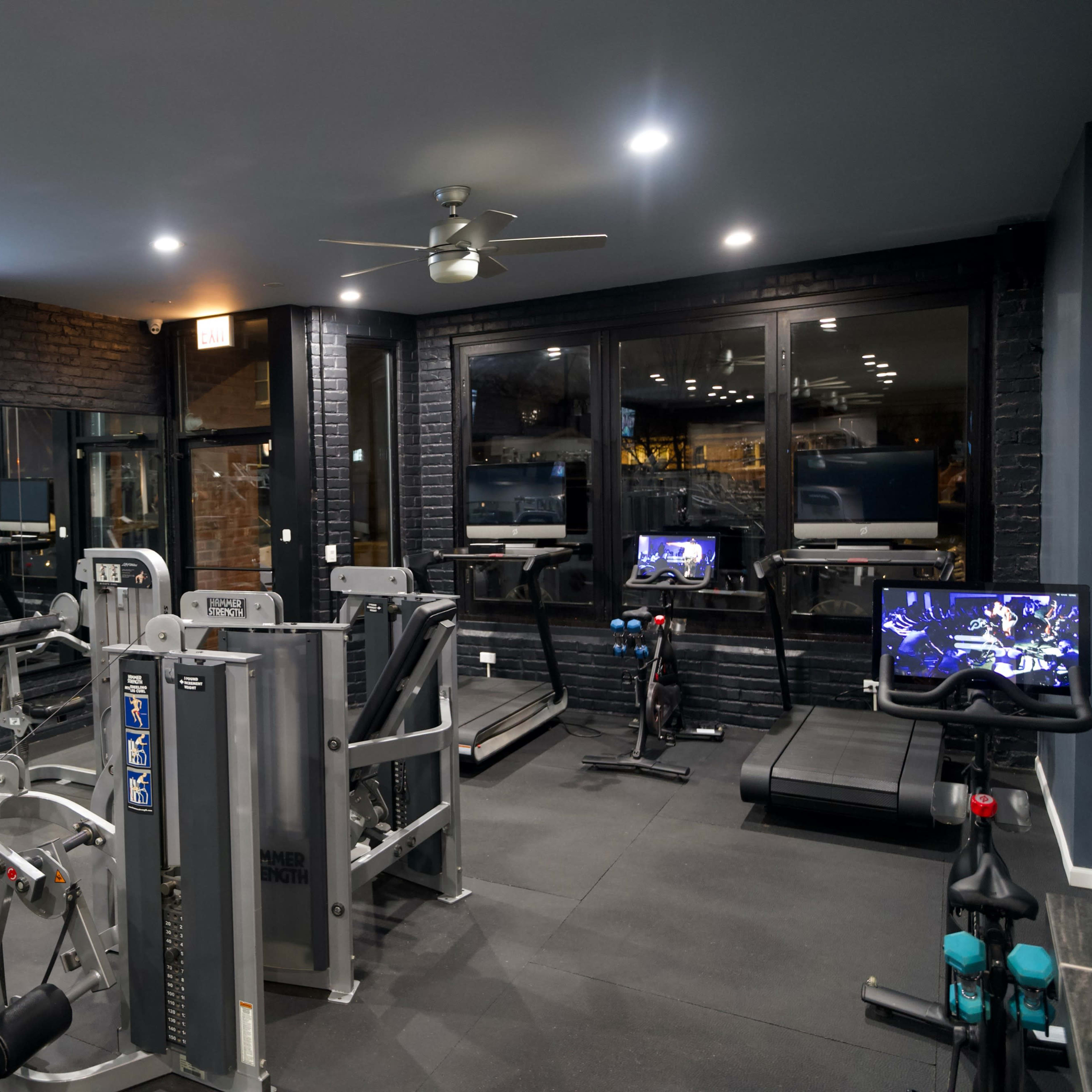 A well-equipped gym with various exercise machines, including treadmills and weight machines, under bright lighting and a ceiling fan.
