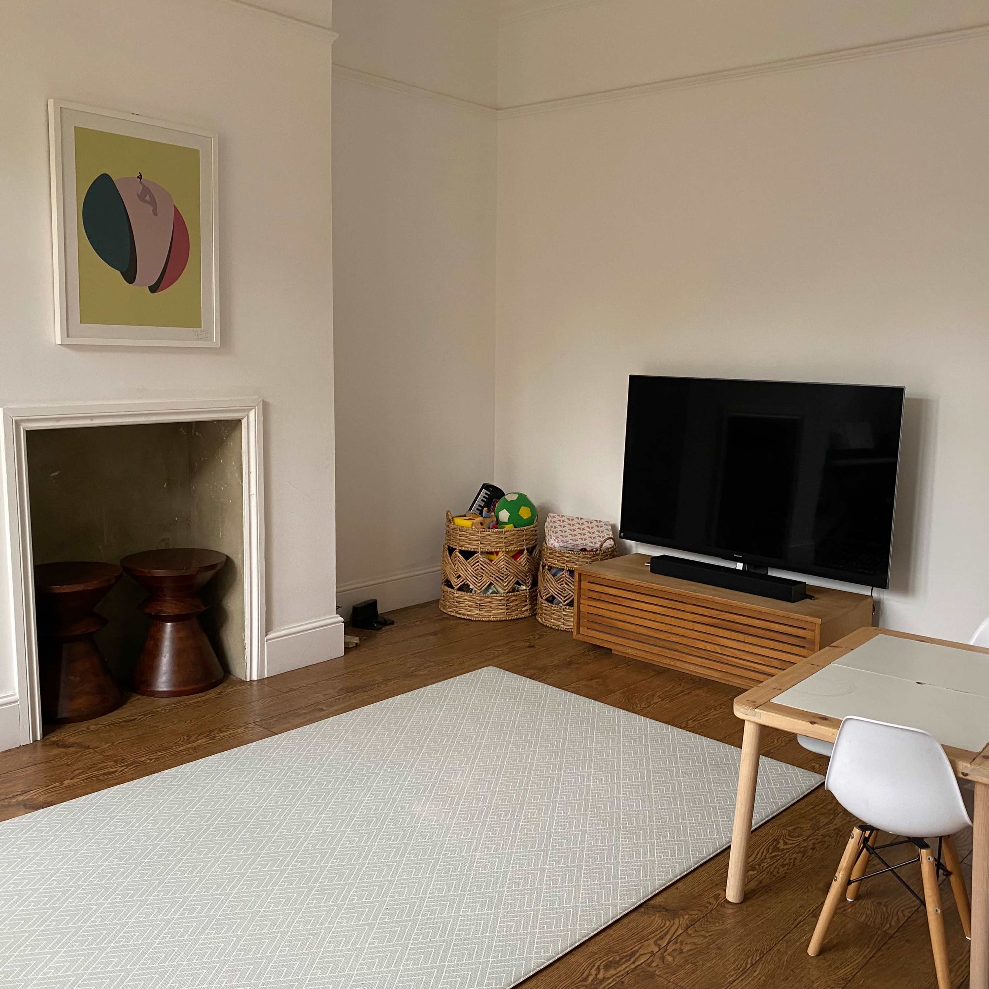 A minimalist living room features a television stand, a small wooden table with a chair, and a patterned area rug on a wooden floor.