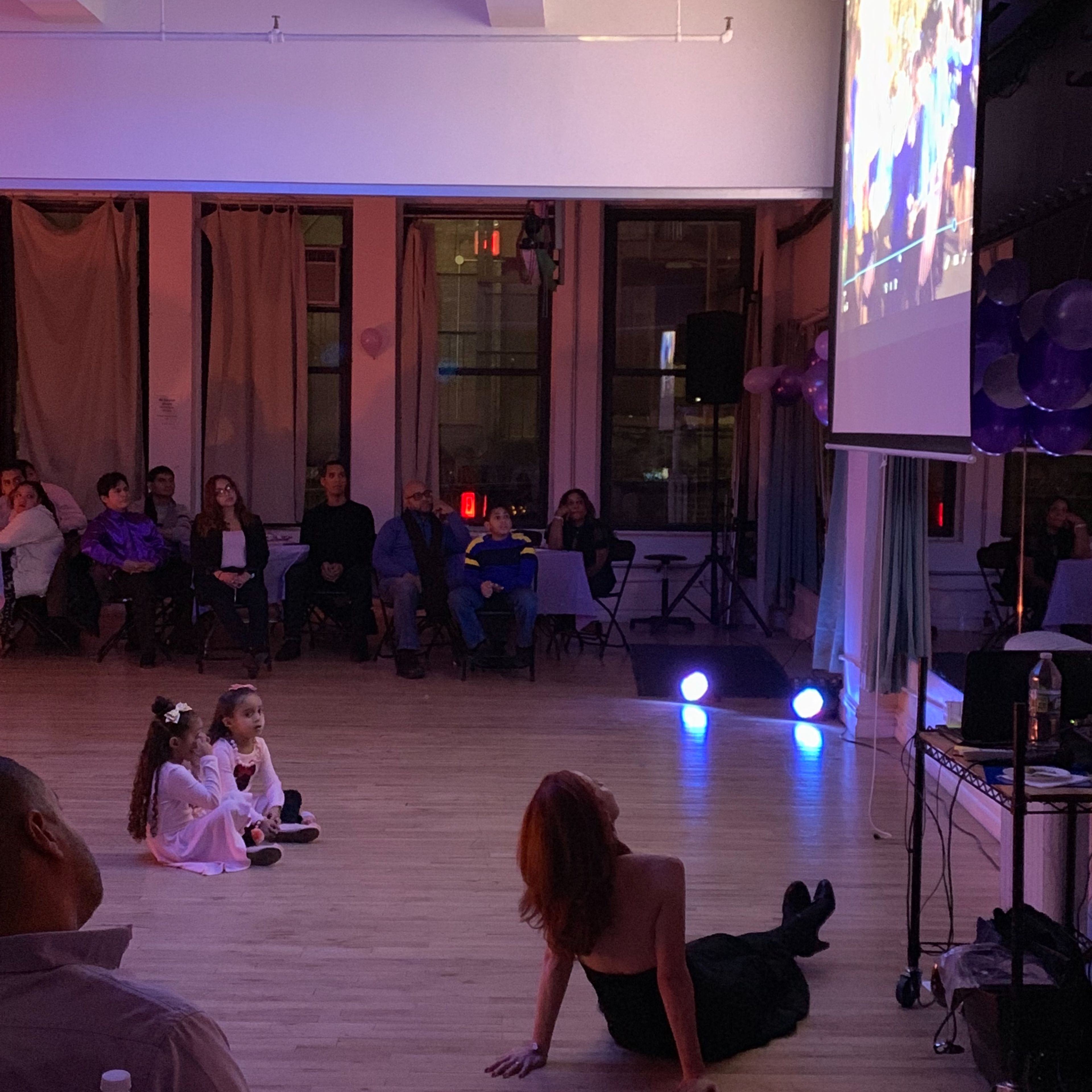 Two young girls sit on the floor watching a video projected on a screen while an audience observes from seated positions.