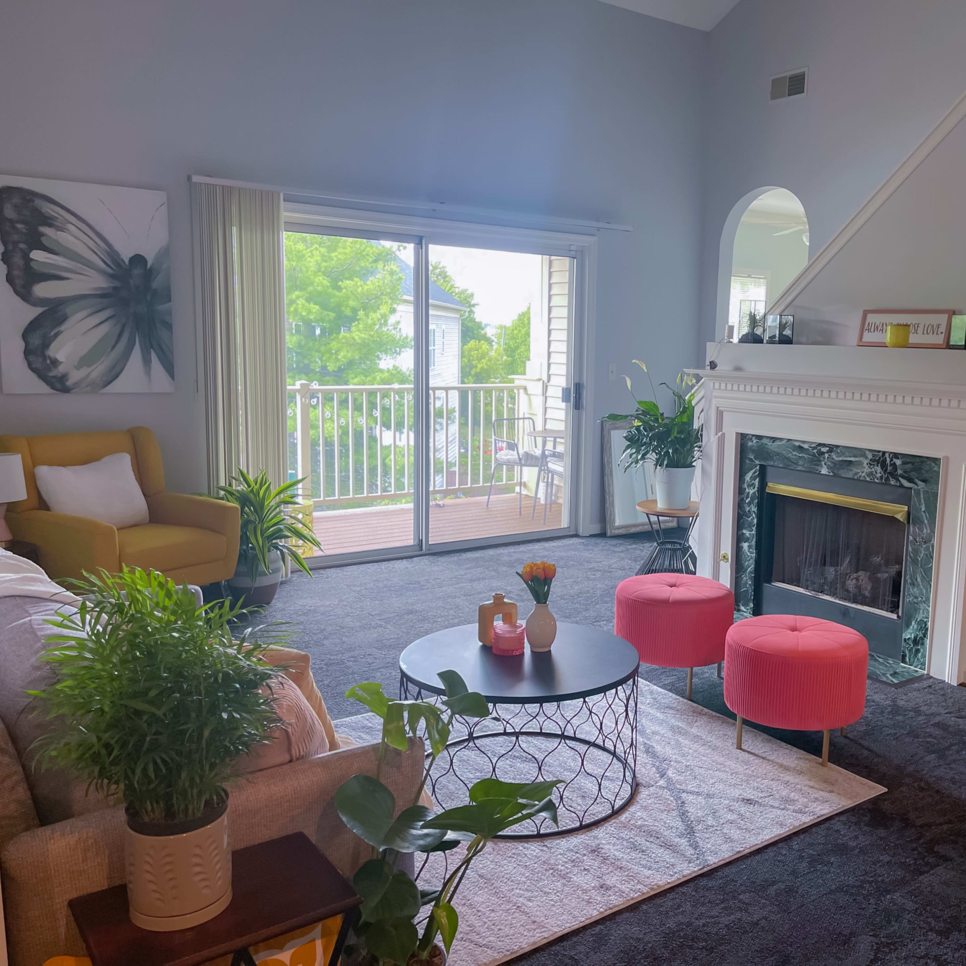 The living room features a butterfly artwork on the wall, a yellow armchair, a beige sofa, a round coffee table, and pink ottomans, with large windows offering a view of a balcony and greenery outside.