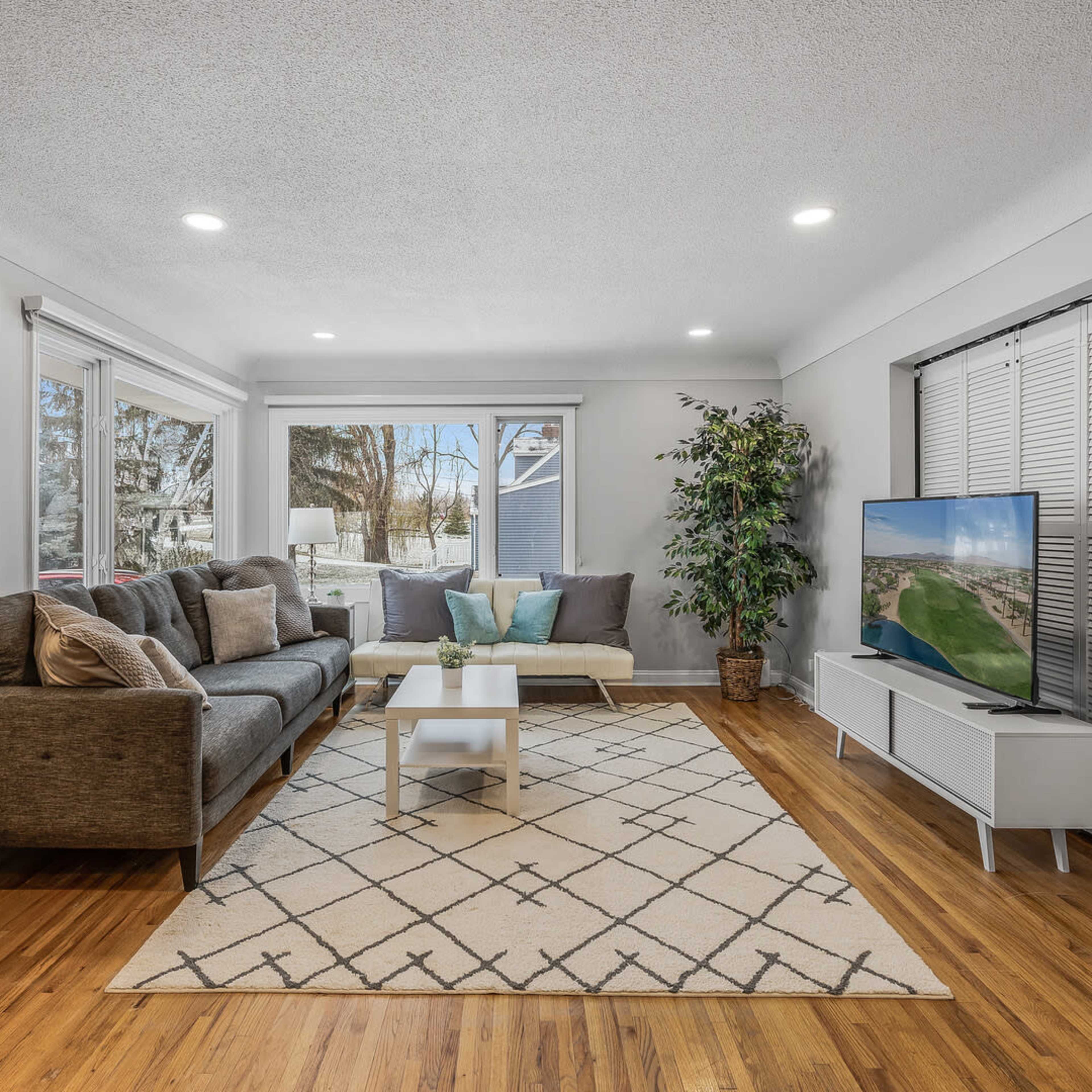 A modern living room with a brown sectional sofa, a coffee table on a geometric patterned rug, and a large television set against the wall, with windows allowing natural light to enter.