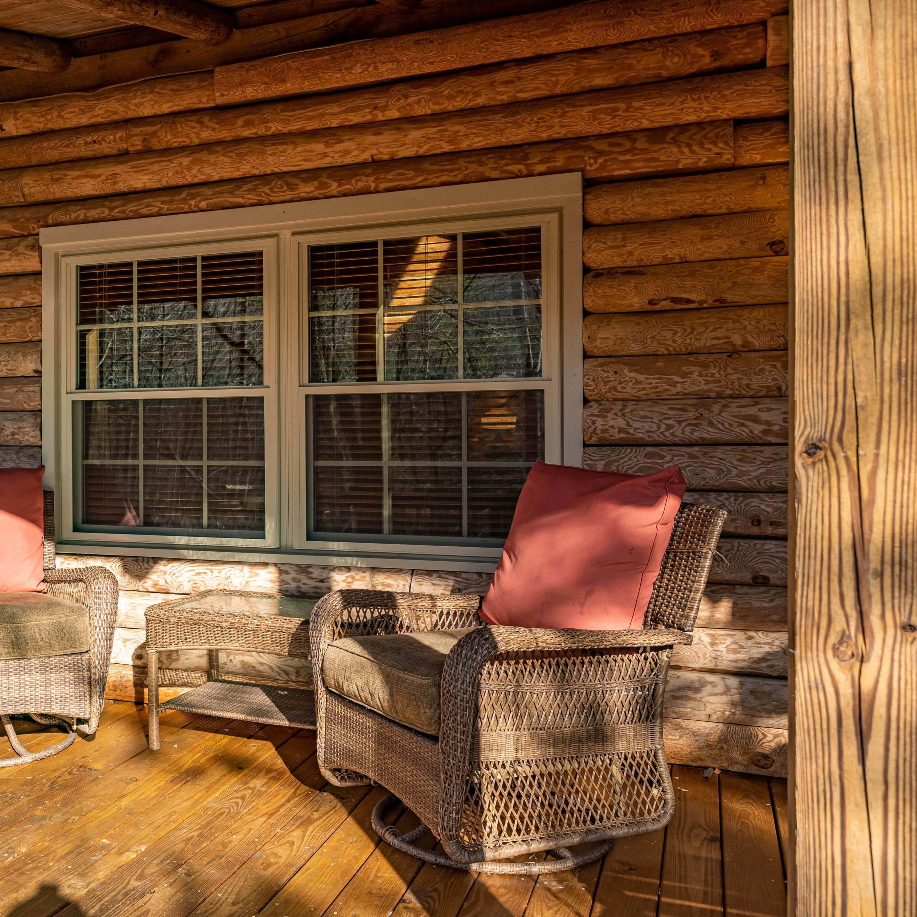 Two wicker chairs with orange cushions are arranged on a wooden porch in front of a log cabin.