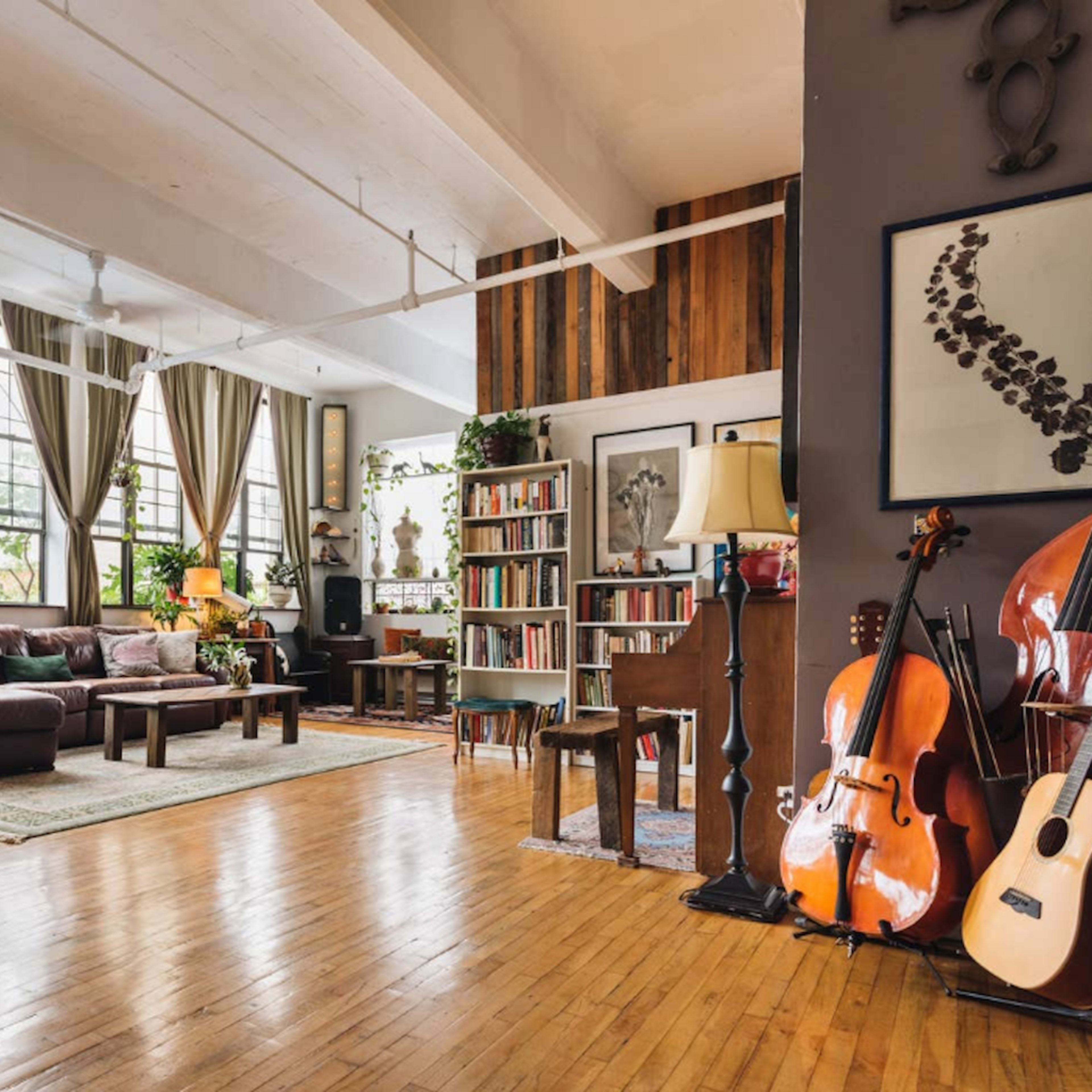 A spacious living area with large windows, hardwood floors, a brown leather sofa, plants, a bookshelf, and various musical instruments.