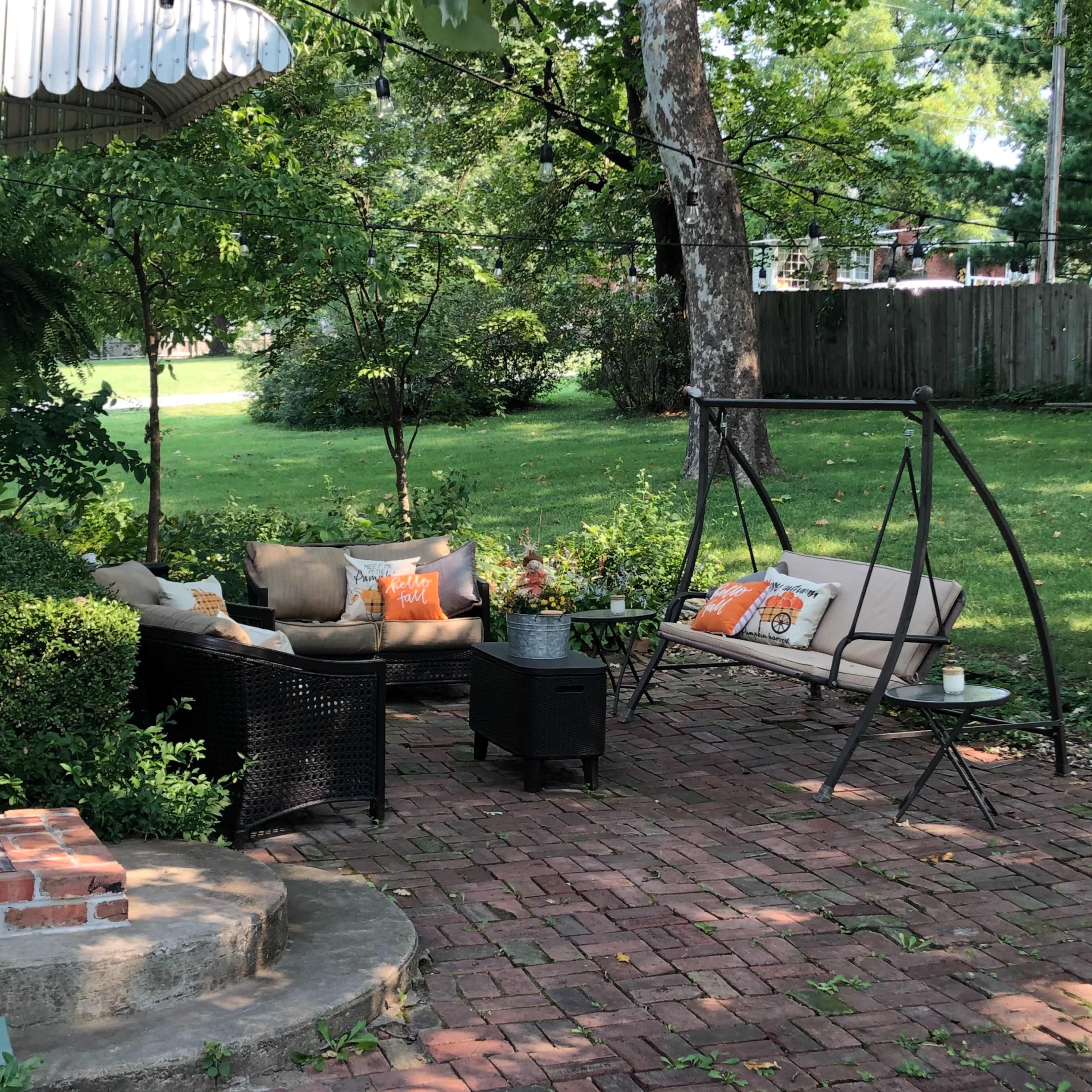 A cozy outdoor seating area features a swing chair and a black wicker sofa surrounded by greenery and brick paving.