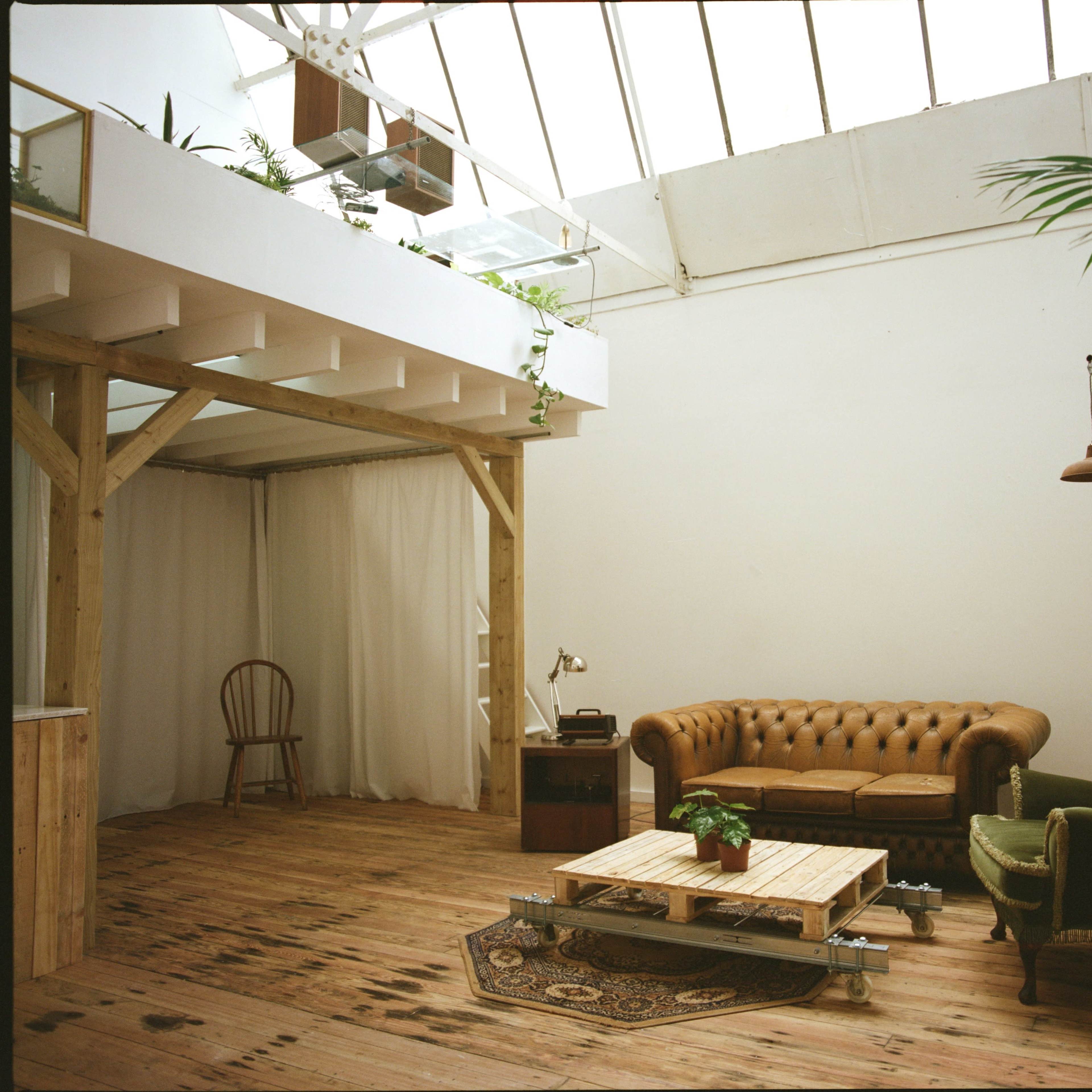 The scene depicts a spacious, well-lit room featuring a wooden floor, a brown leather chesterfield sofa, a green armchair, and a wooden coffee table, with large windows allowing natural light to fill the space.