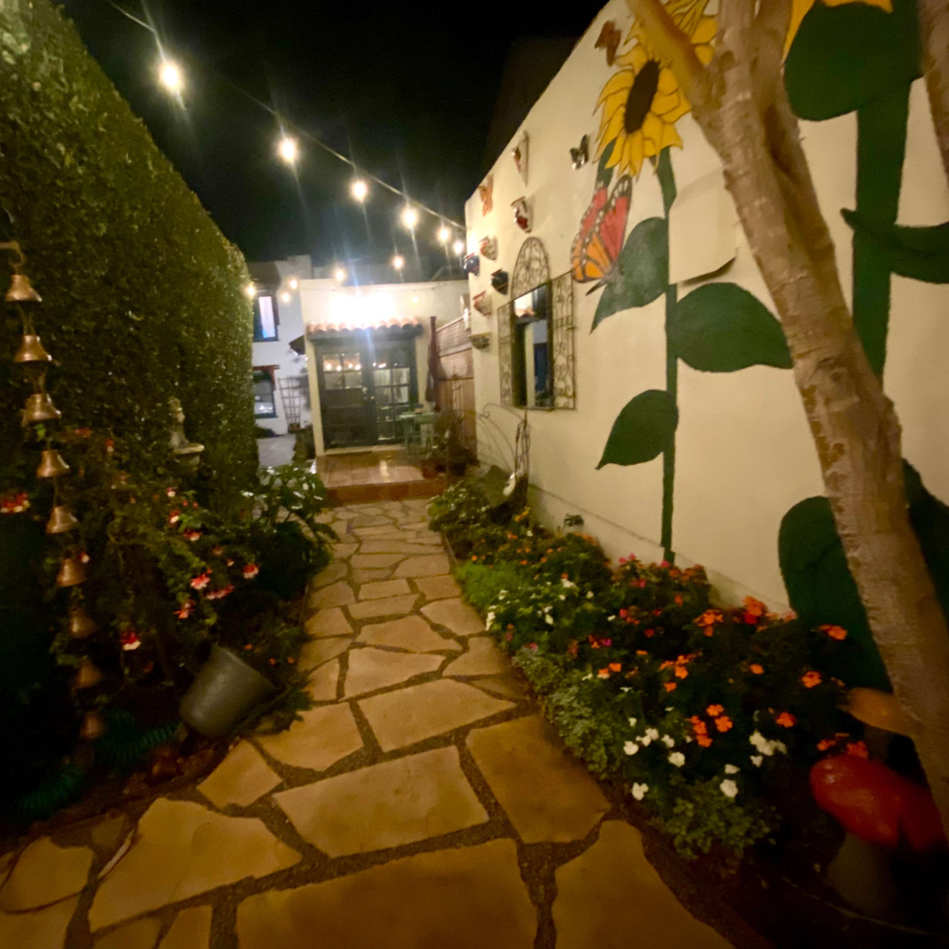 A stone pathway illuminated by string lights, surrounded by decorative plants and a mural of sunflowers on a wall.