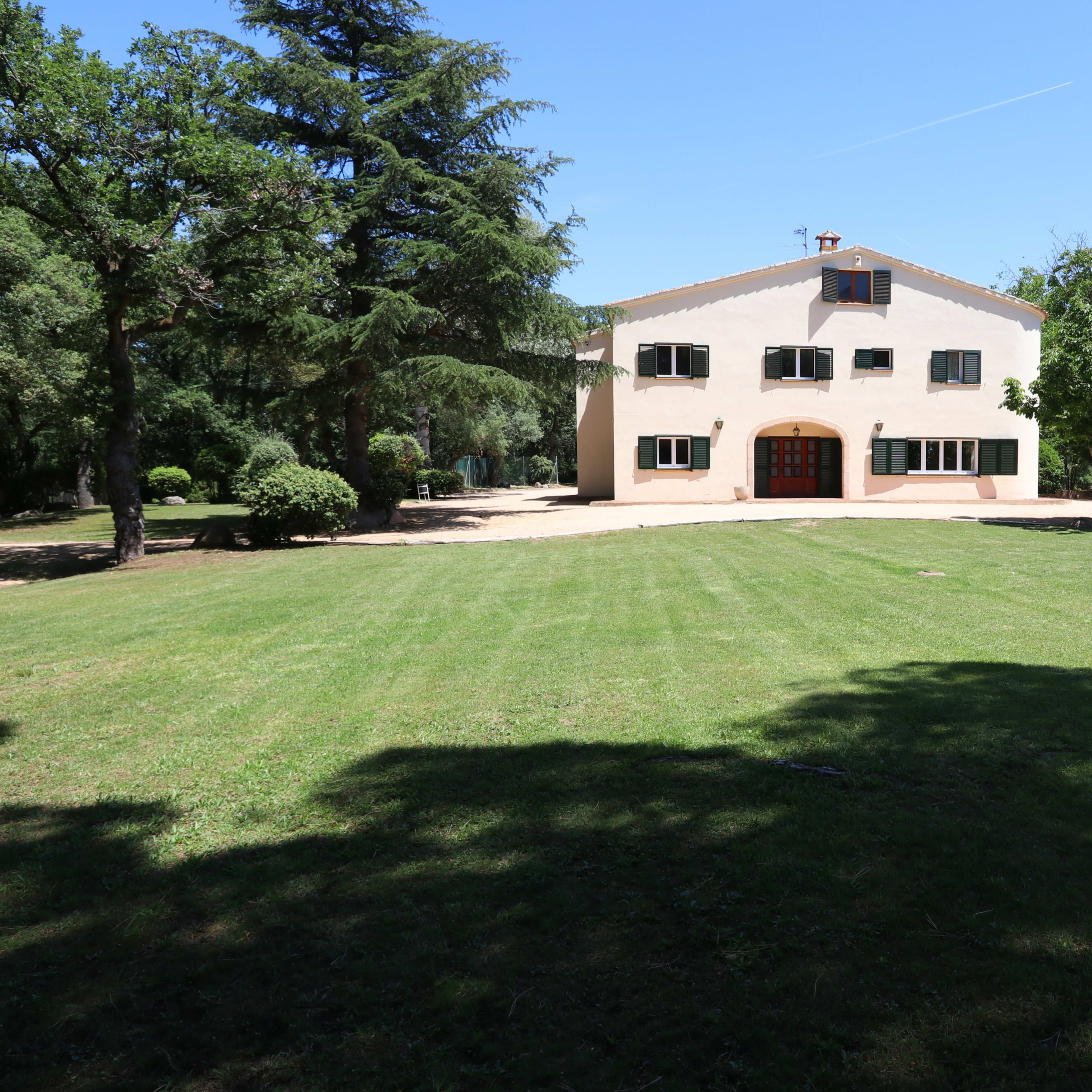 A large, two-story house with multiple windows stands in a grassy yard surrounded by tall trees.