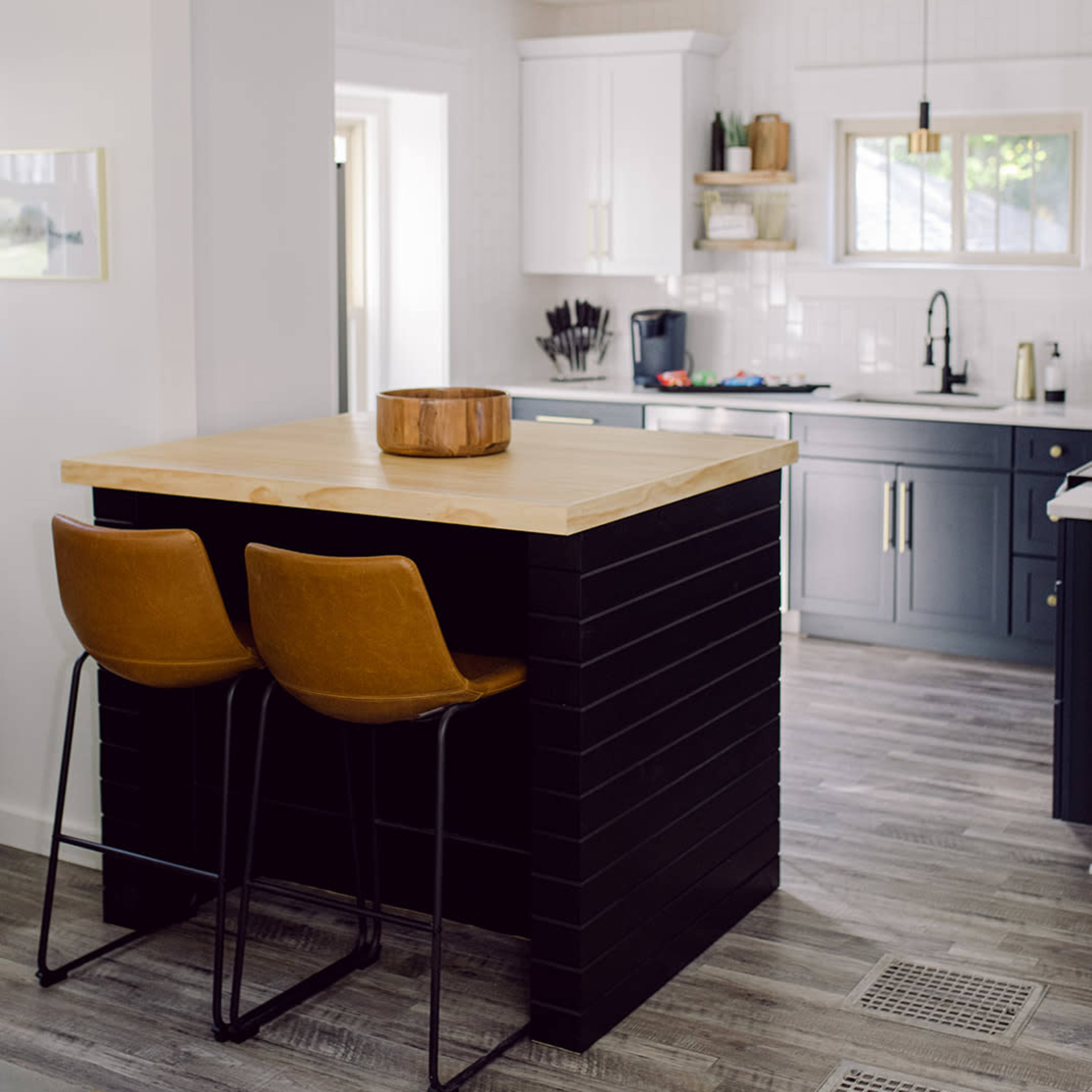 A modern kitchen featuring a wooden countertop with two brown bar stools, a dark cabinetry unit, and white cabinets set against light walls.