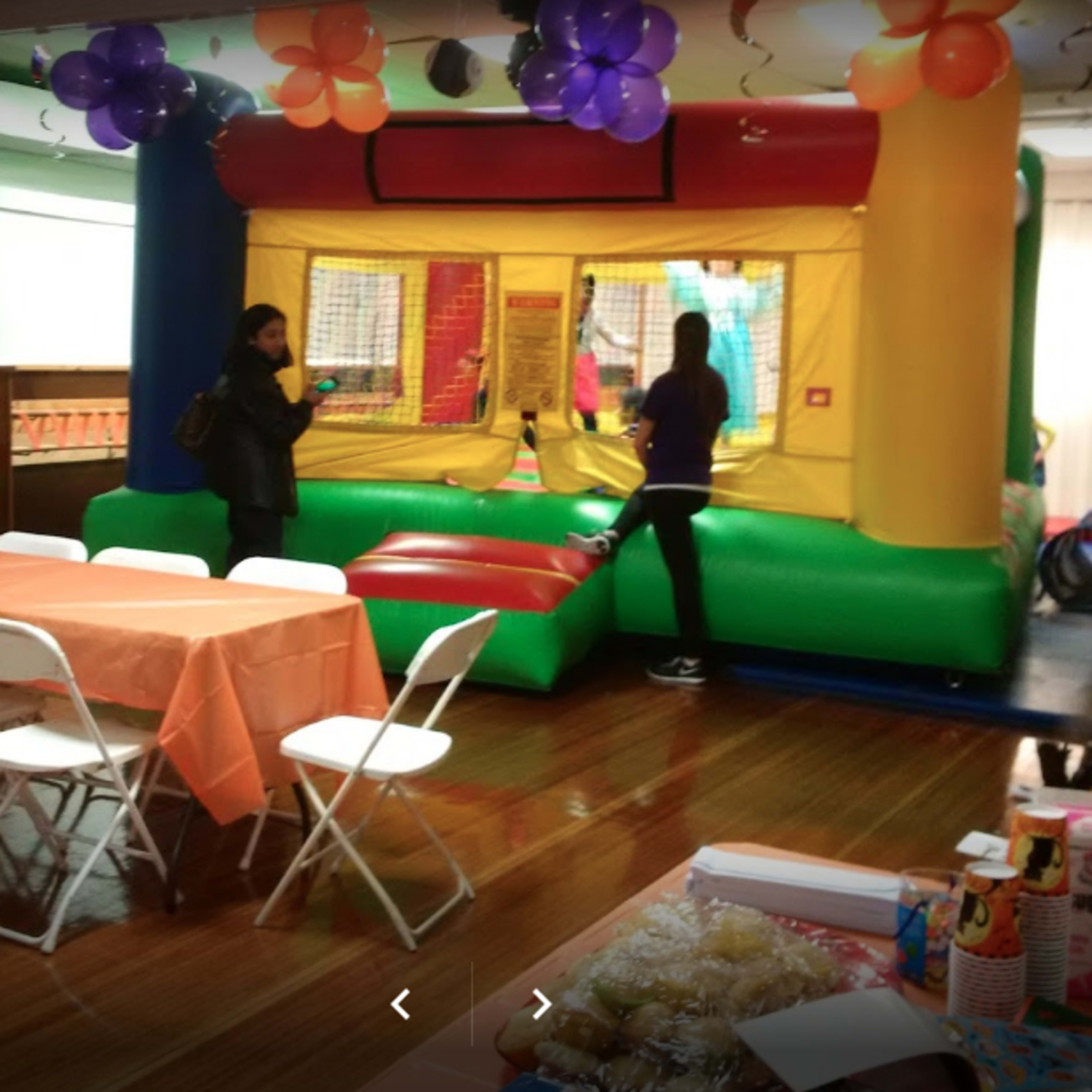 Two women interact with a colorful inflatable bounce house in a room set up with a table and chairs.