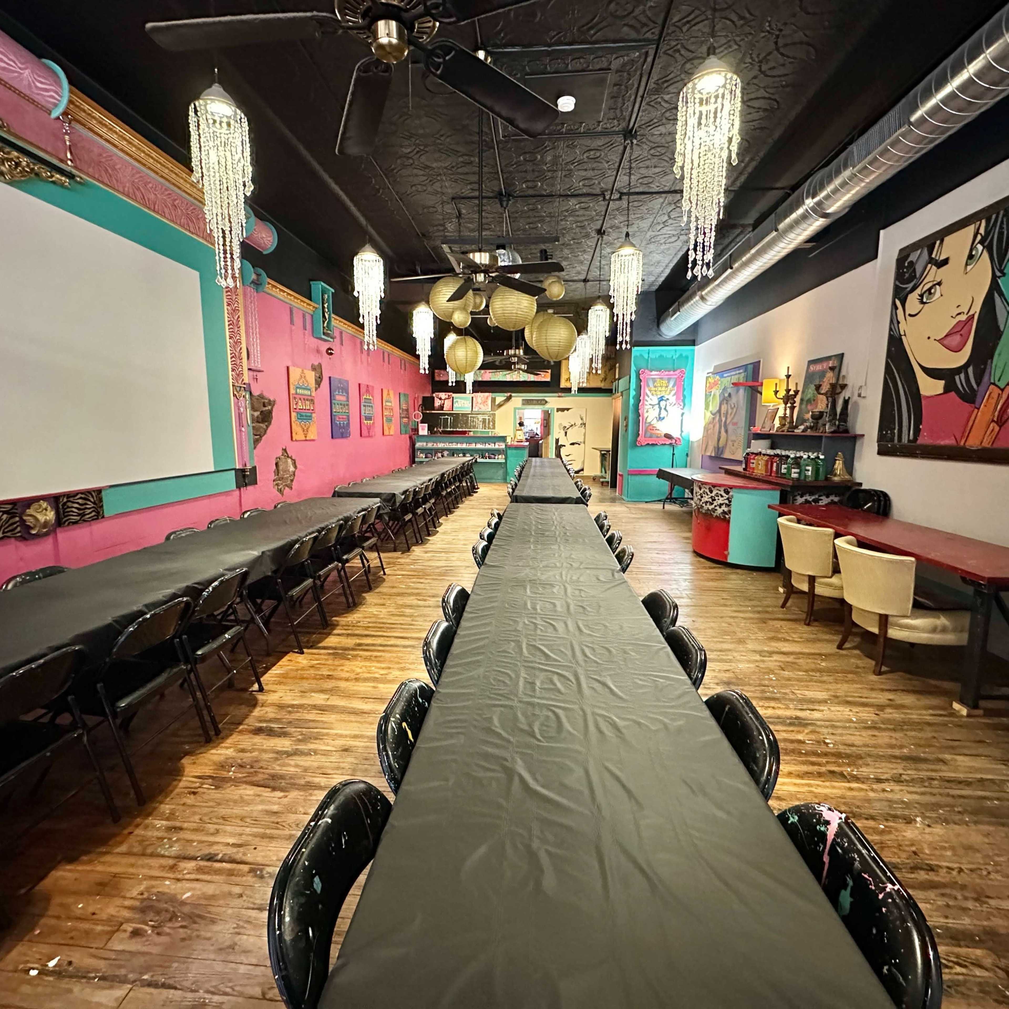 A colorful event space with a long, black-topped table set up in the center, surrounded by chairs, and decorated with vibrant wall art and hanging light fixtures.
