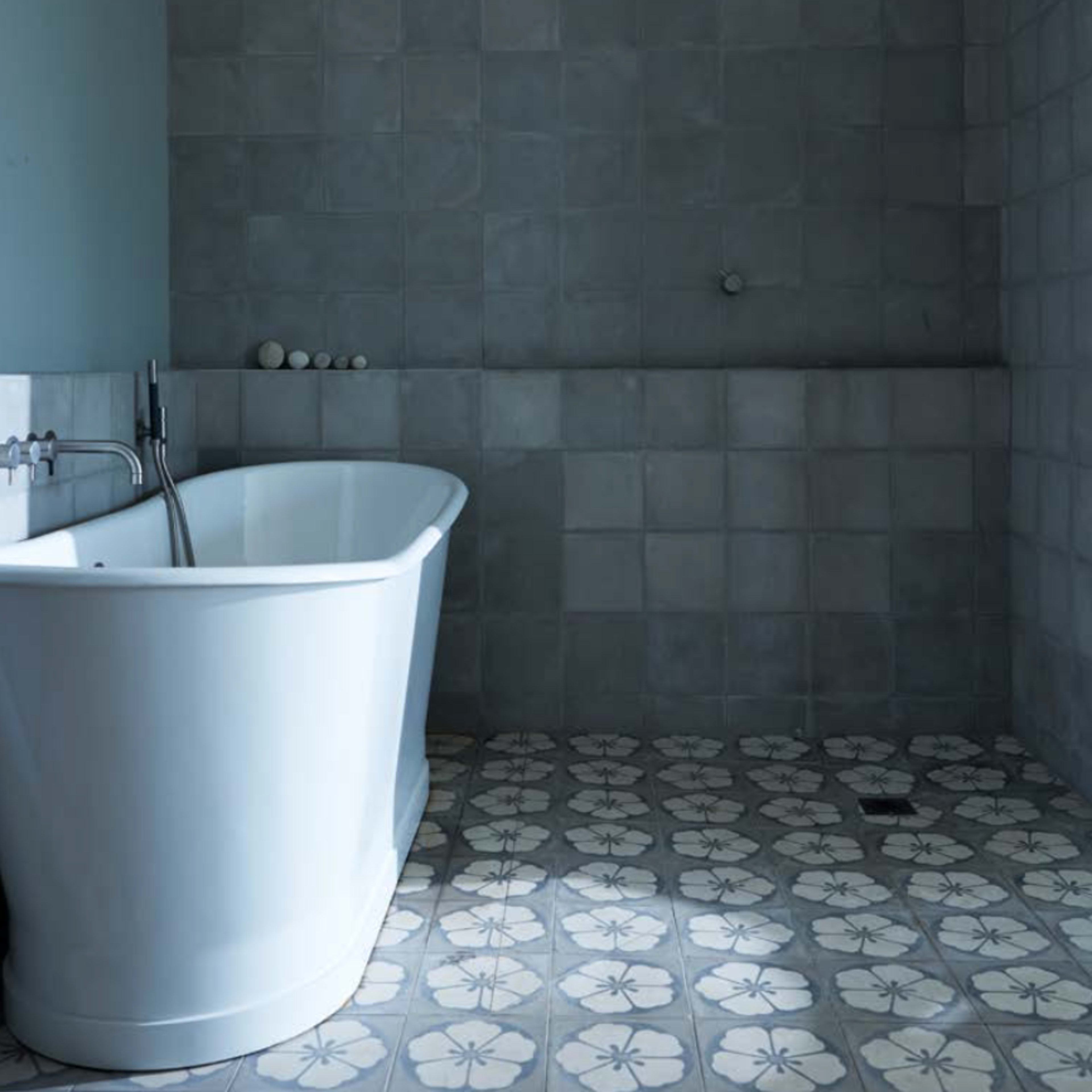 A modern bathroom featuring a white freestanding bathtub against a backdrop of gray tiled walls and patterned floor tiles.
