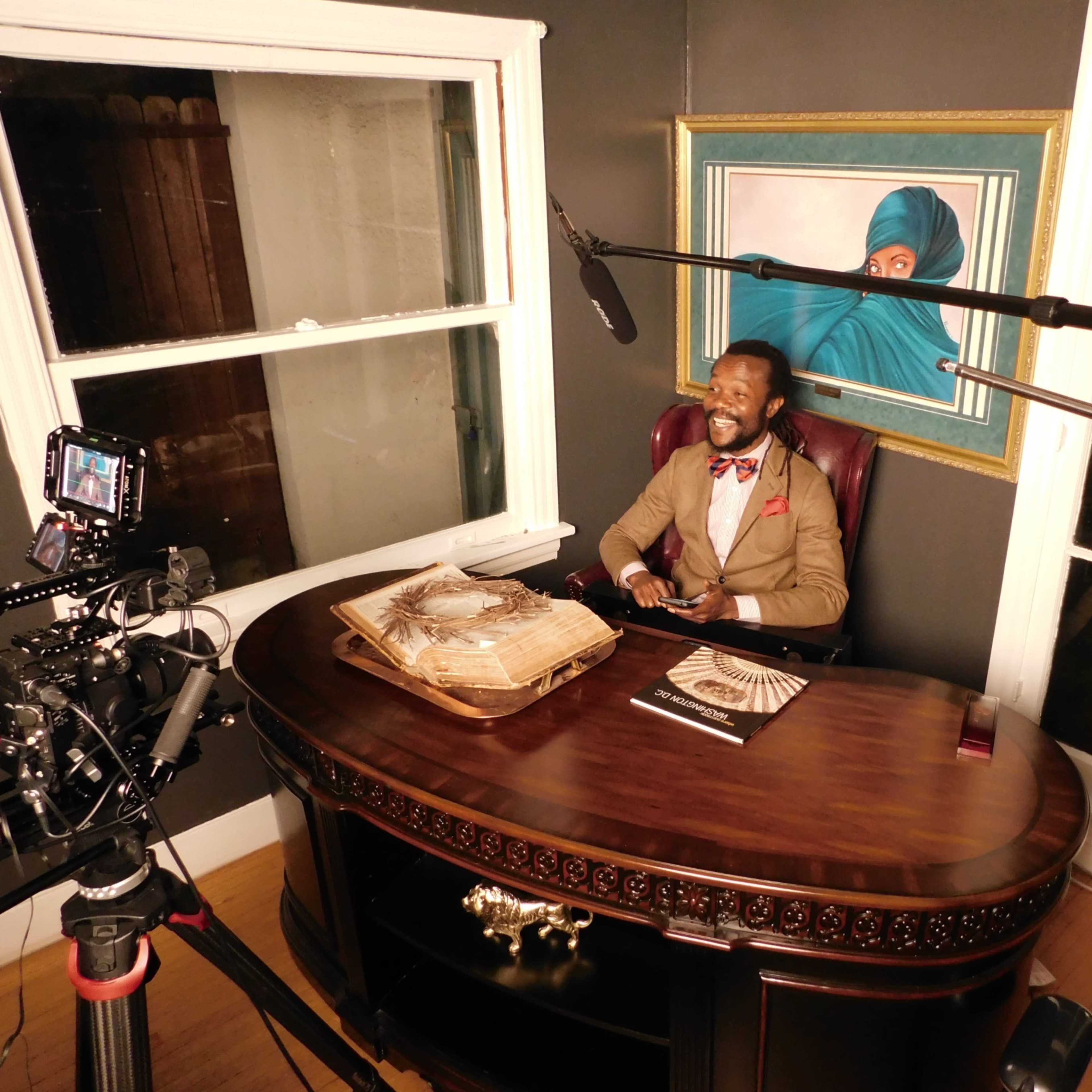 A man sits at a large wooden desk with a camera and microphones set up for recording in a well-lit room featuring a painting on the wall.
