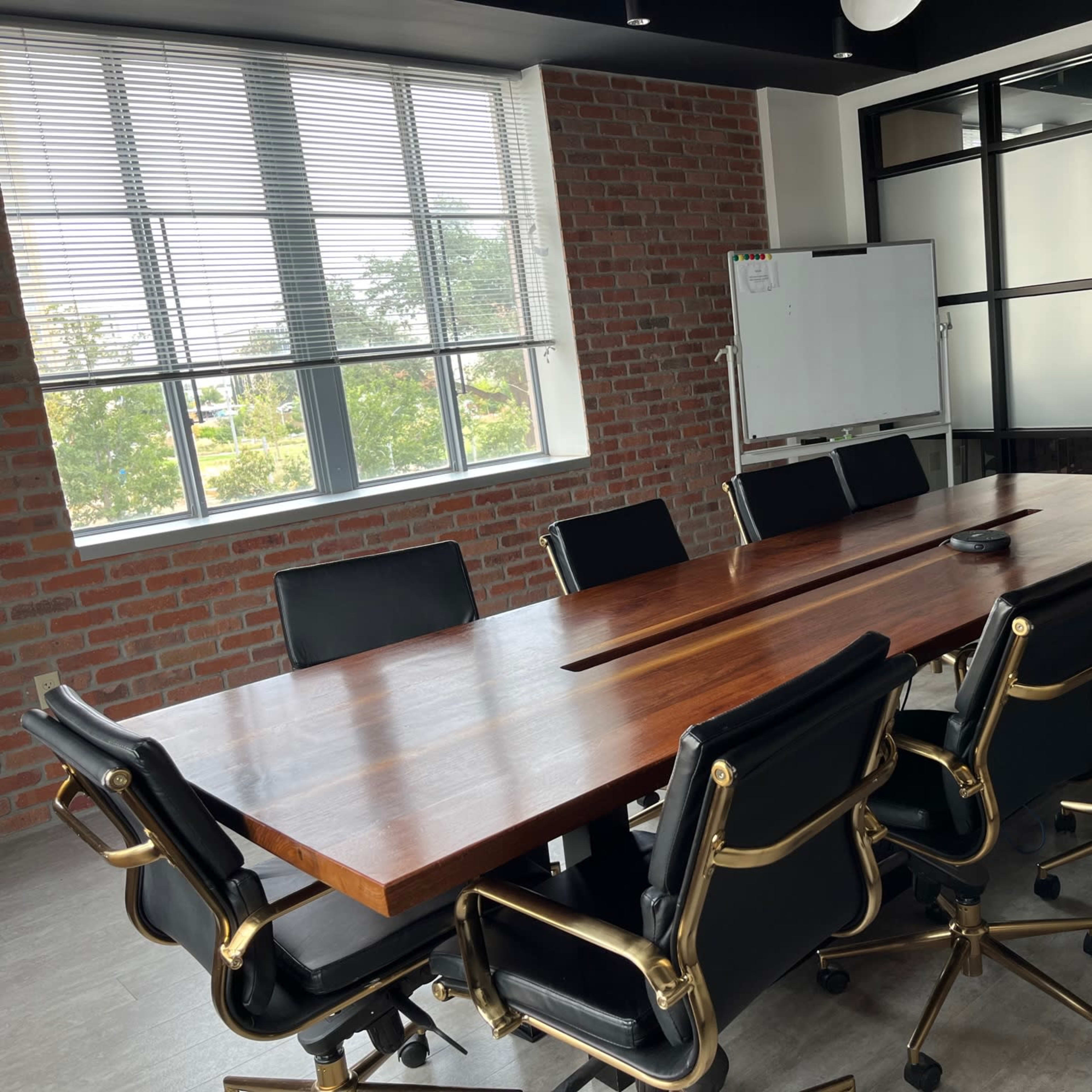A large wooden conference table is surrounded by black office chairs in a bright meeting room with brick walls and large windows.