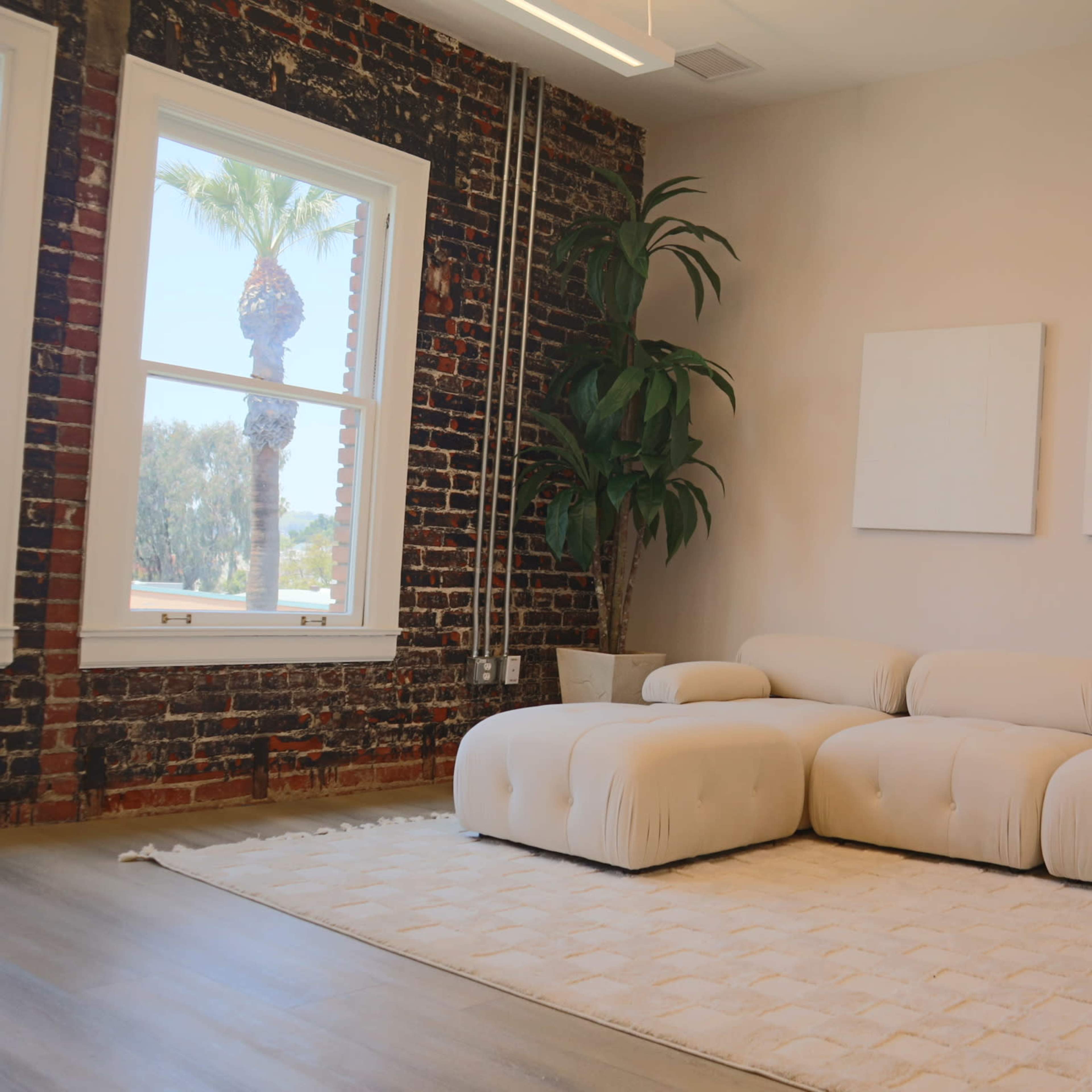 A modern living area featuring a light-colored sectional sofa, two blank white canvas frames on the wall, and a large window with a view of palm trees, contrasted against a red brick wall.