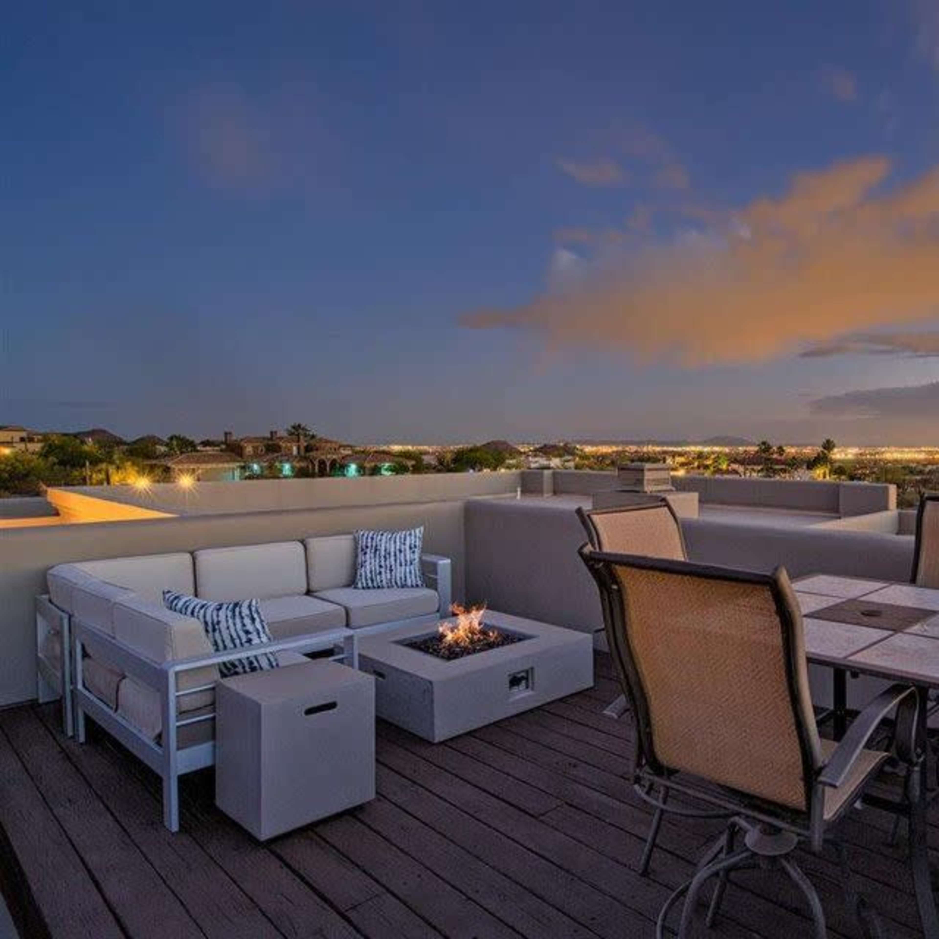 A rooftop patio with seating arrangements, a fire pit, and a view of the city skyline under a twilight sky.