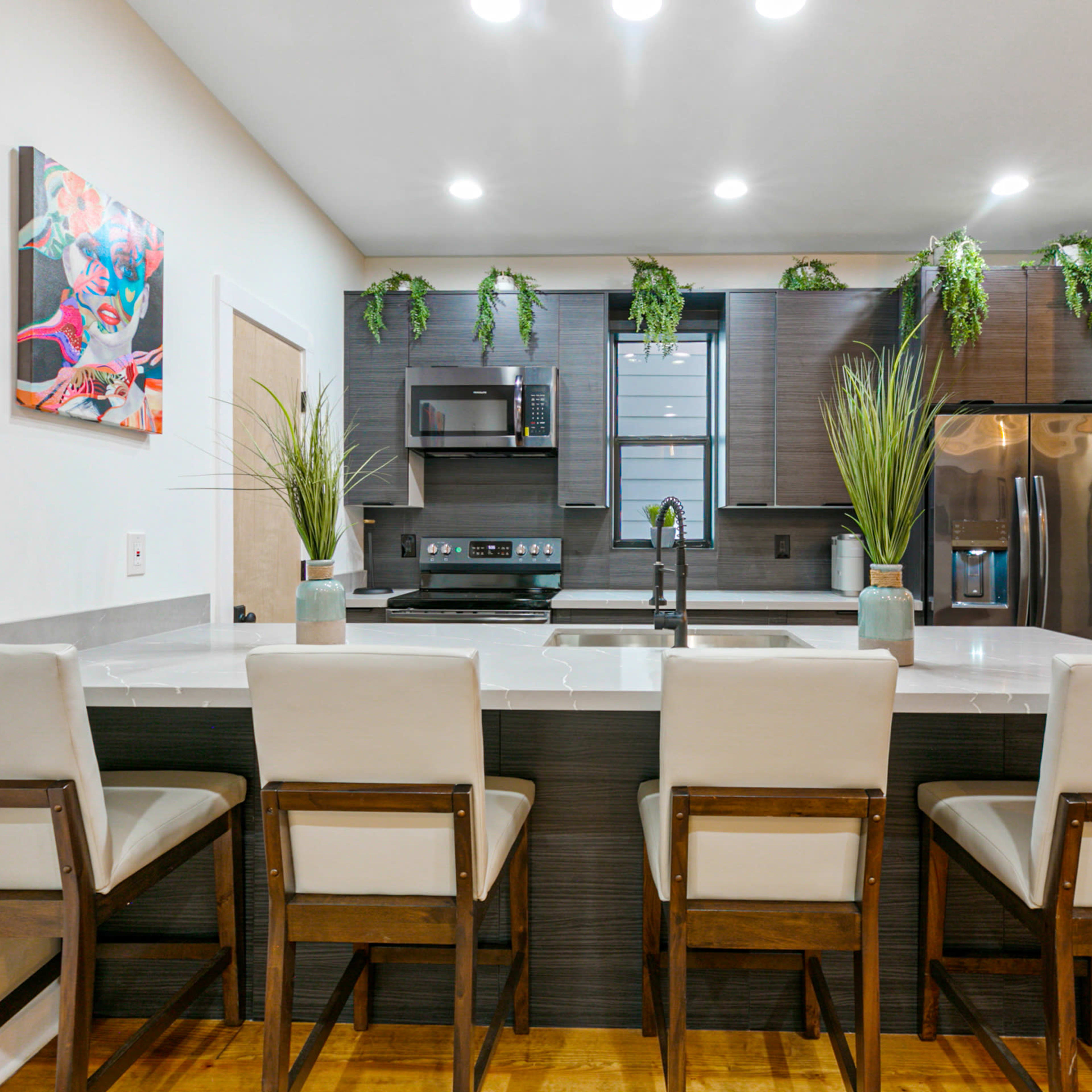 A modern kitchen features a counter with four upholstered stools, stainless steel appliances, and decorative greenery.