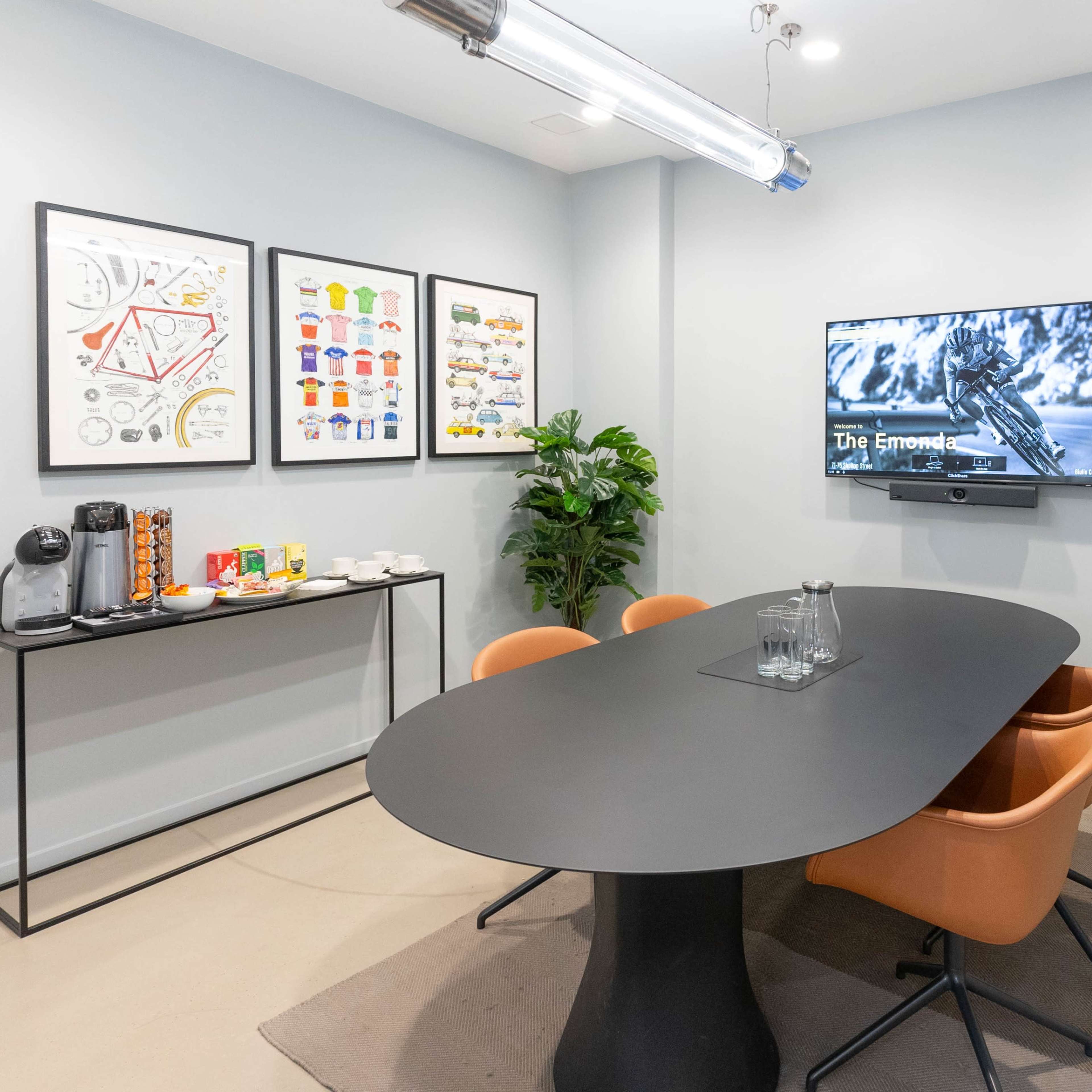 A modern meeting room features a black oval table surrounded by four orange chairs, a wall-mounted TV, plants, and framed art.