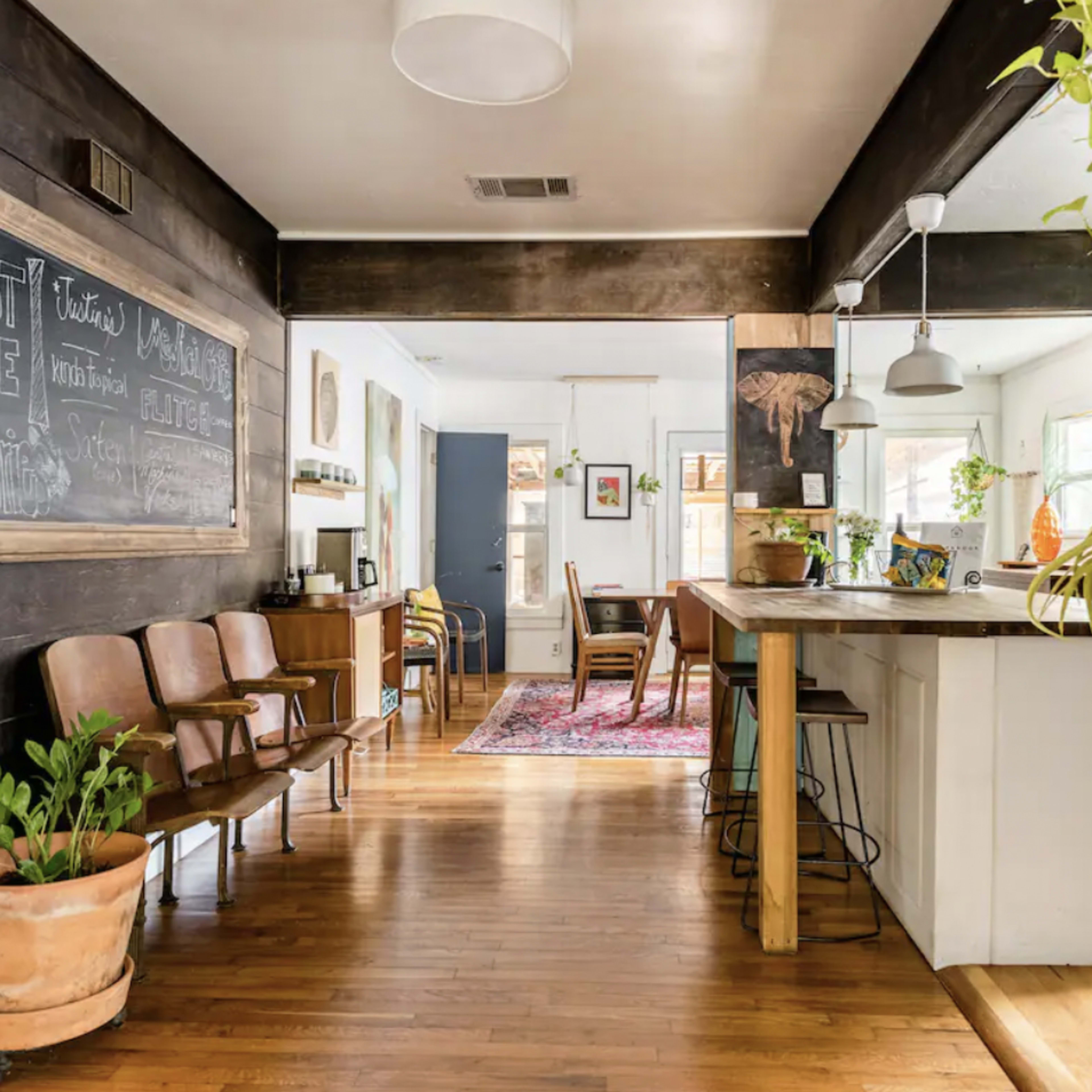A spacious room with wooden flooring, featuring a hallway leading to a dining area and a kitchen, lined with plants and wooden furniture.