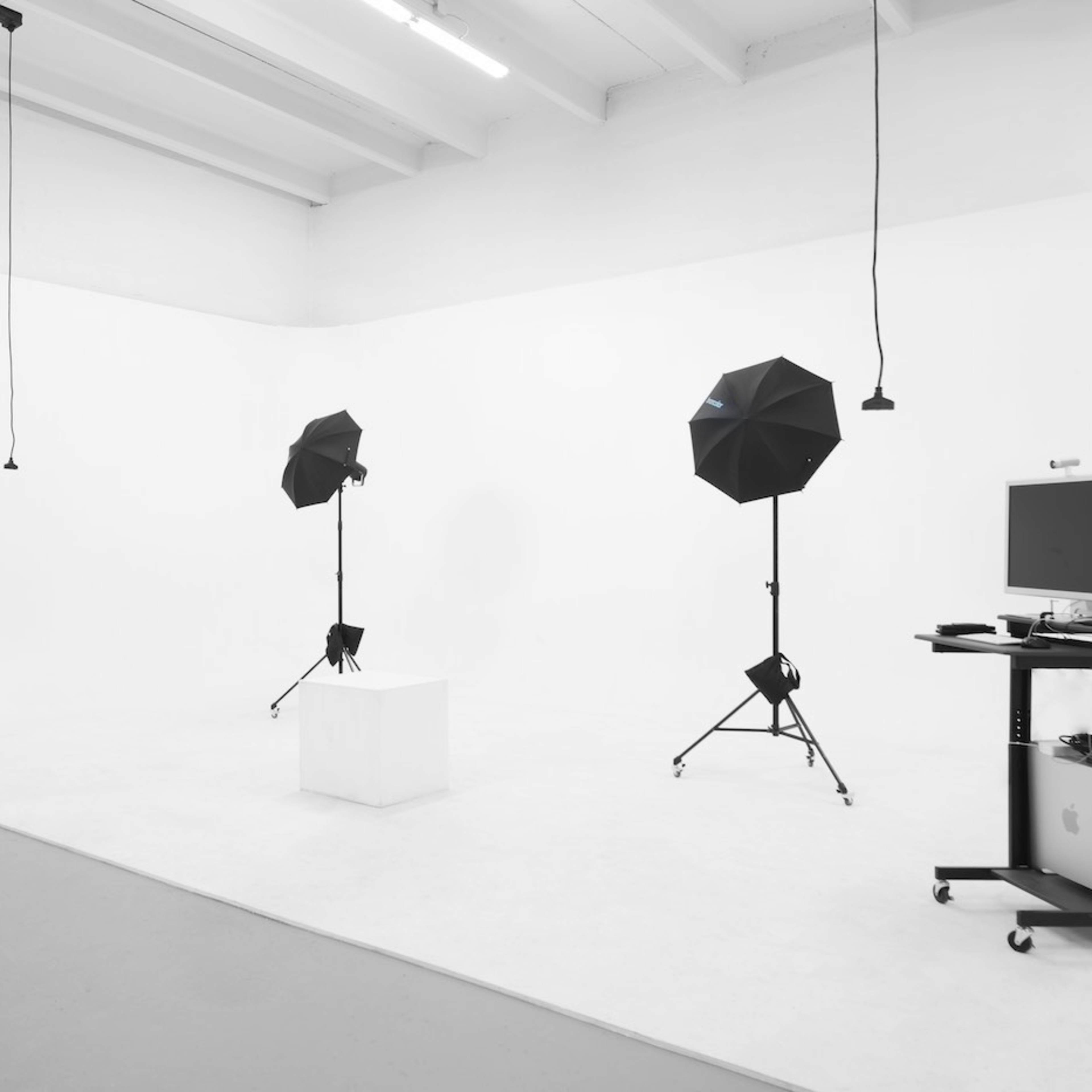 A minimalist photography studio with two black umbrella lights on tripods facing a white backdrop and a small pedestal in the center.