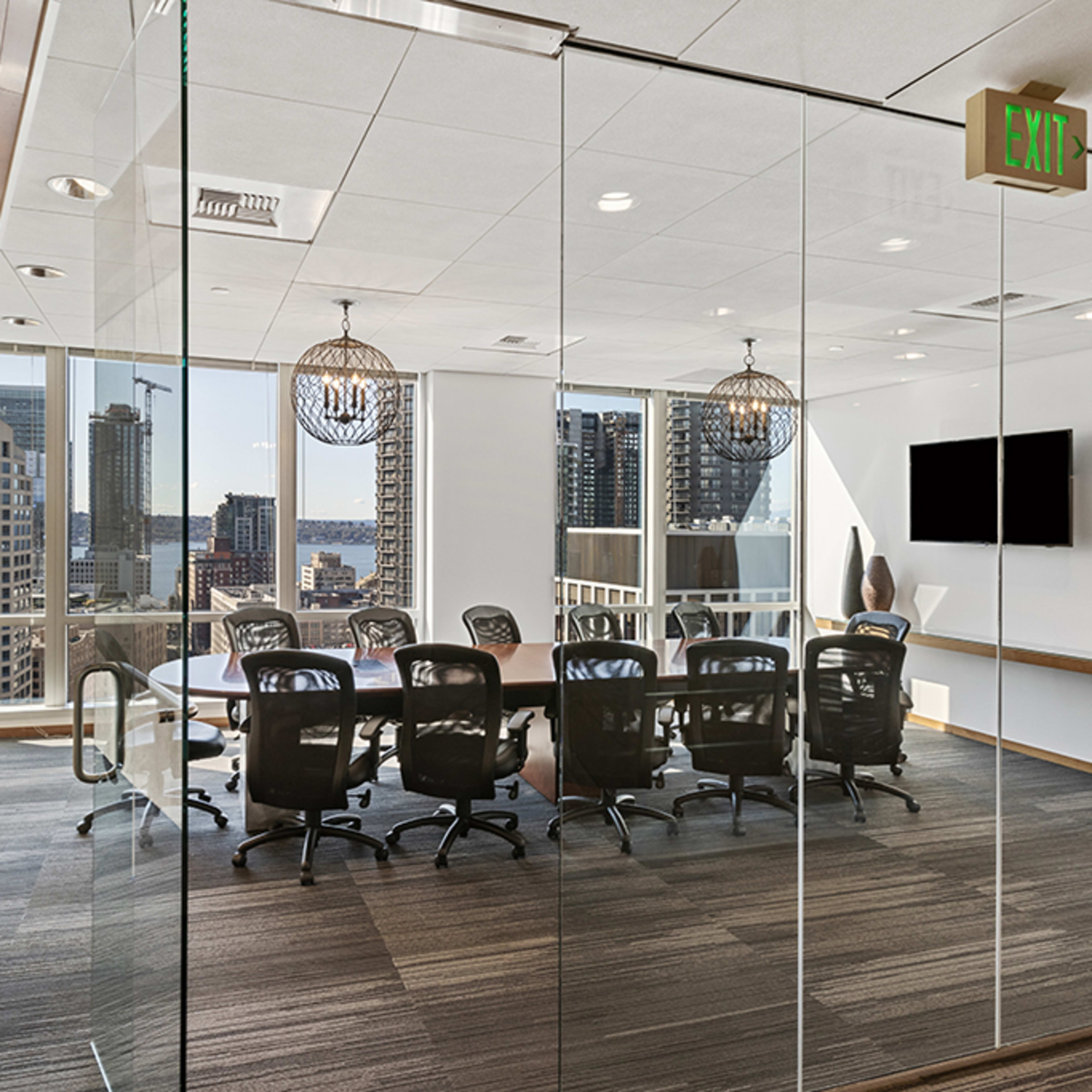 A modern glass-walled conference room features a large table surrounded by black chairs, with a view of a city skyline through the windows.
