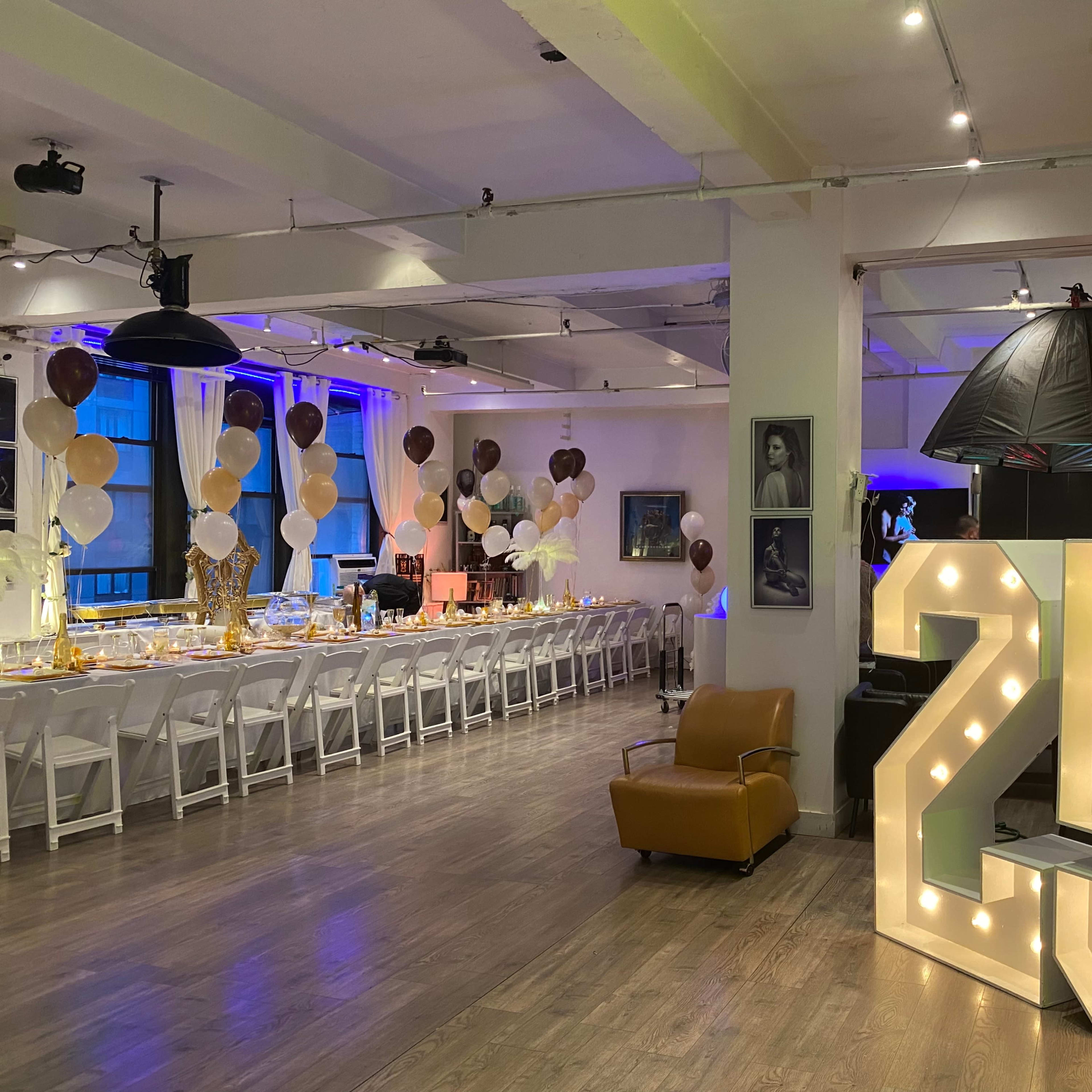 A decorated event space featuring a large illuminated "25," a long dining table set with gold tableware, and balloon arrangements in various colors.