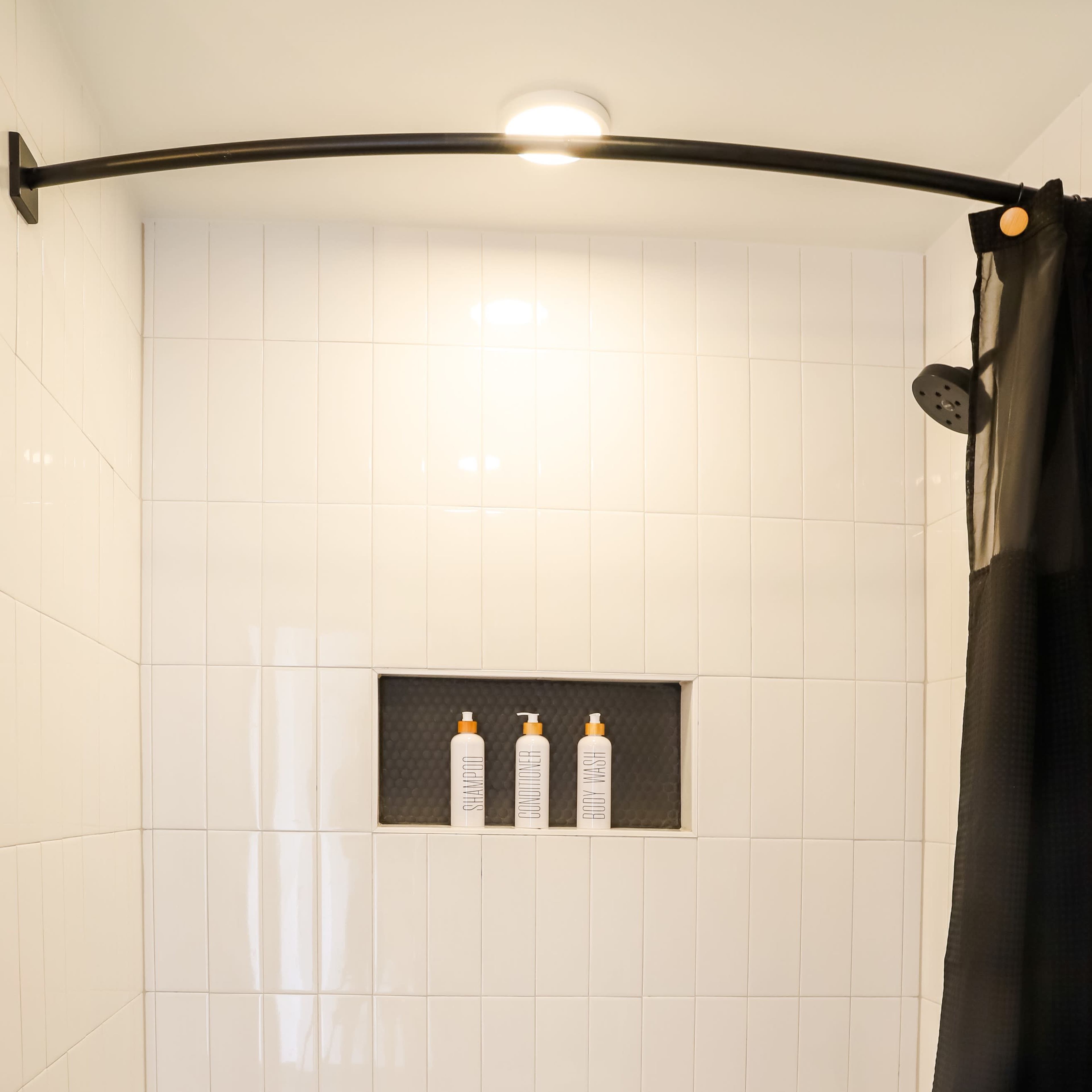 A bathroom with a shower area featuring a black curtain, a curved shower rod, and three bottled products neatly arranged on a shelf in a tiled wall.
