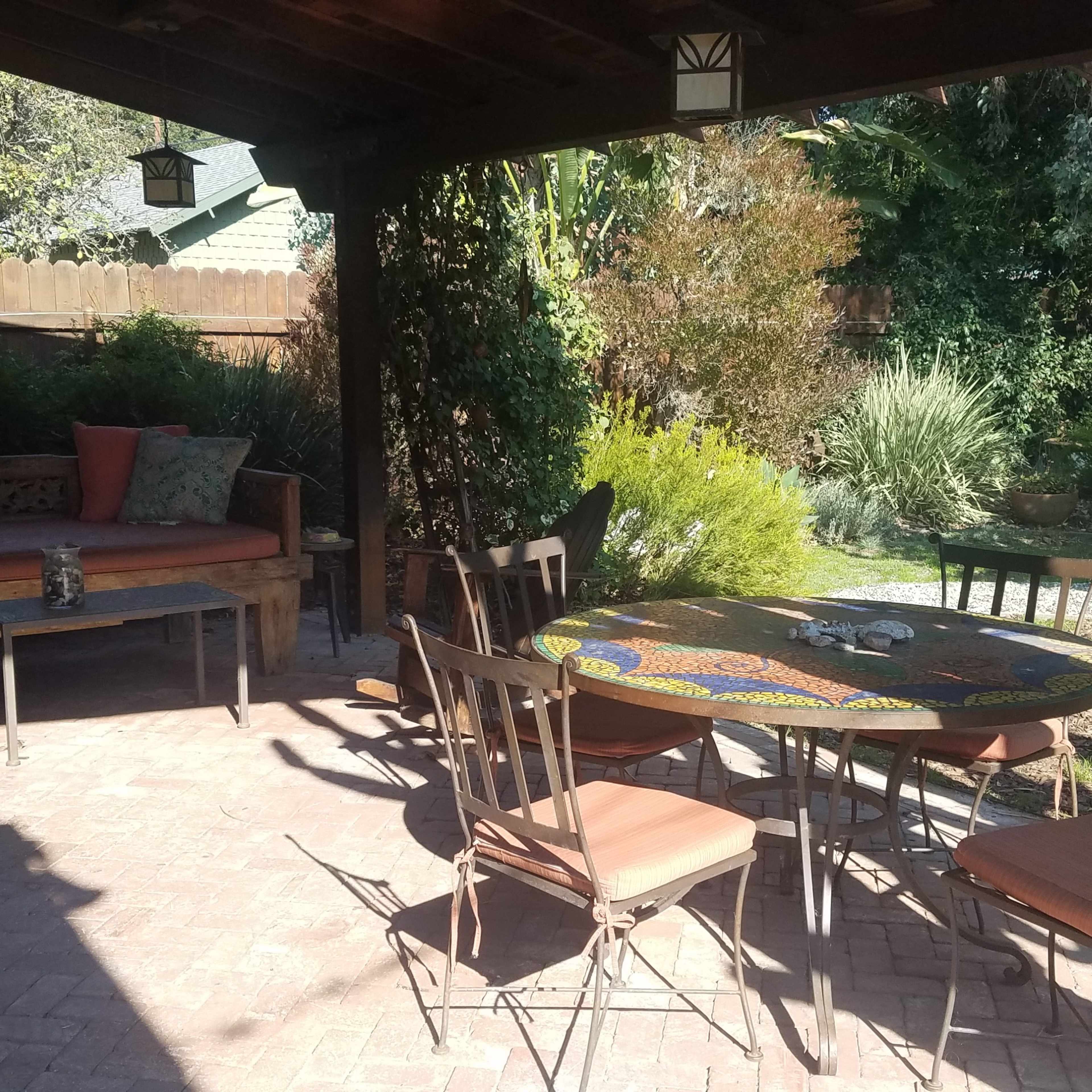 A shaded outdoor patio area with a round table, six chairs, and a wooden bench surrounded by greenery.