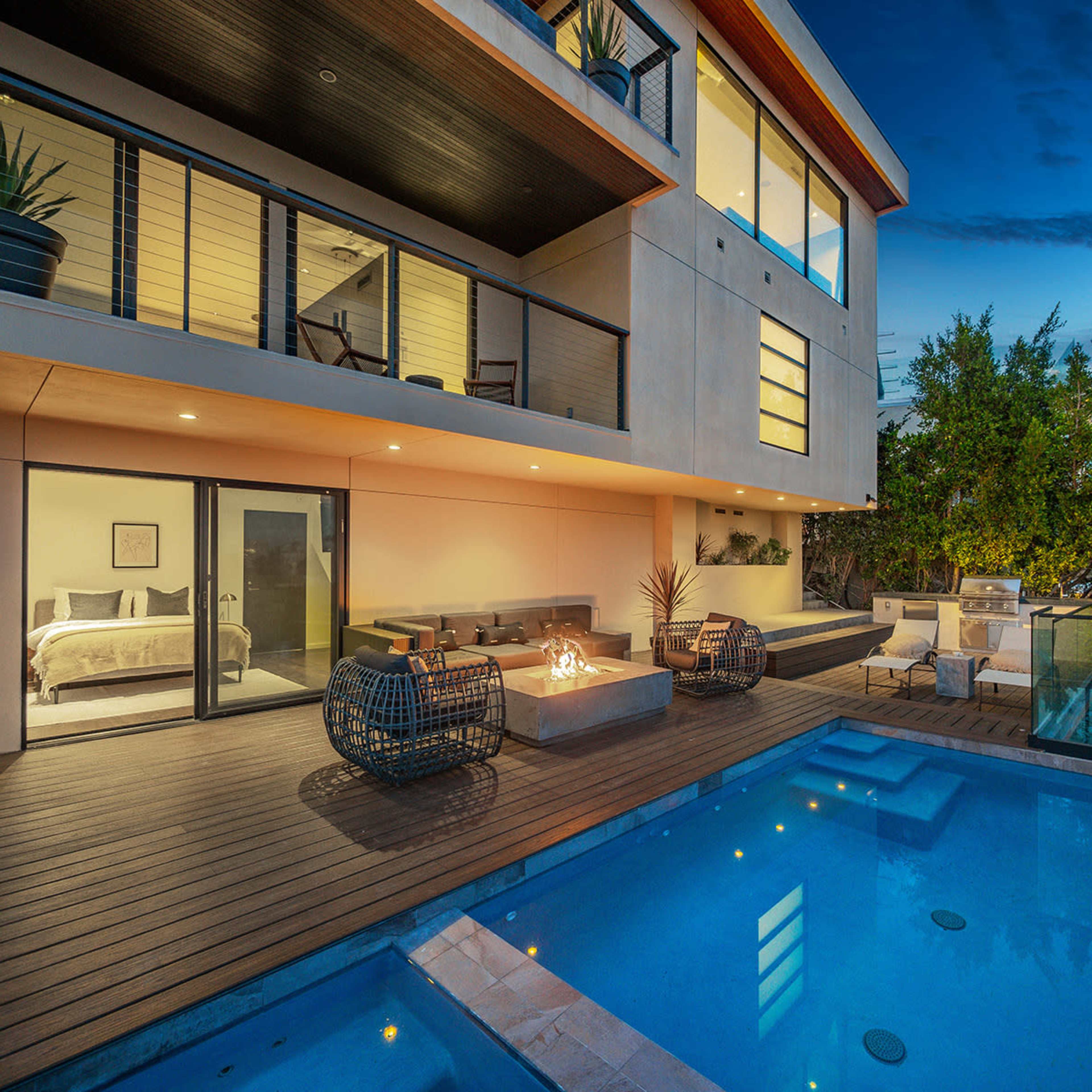 A modern two-story house with large windows overlooking a wooden deck and a swimming pool, featuring outdoor seating and a fire pit.