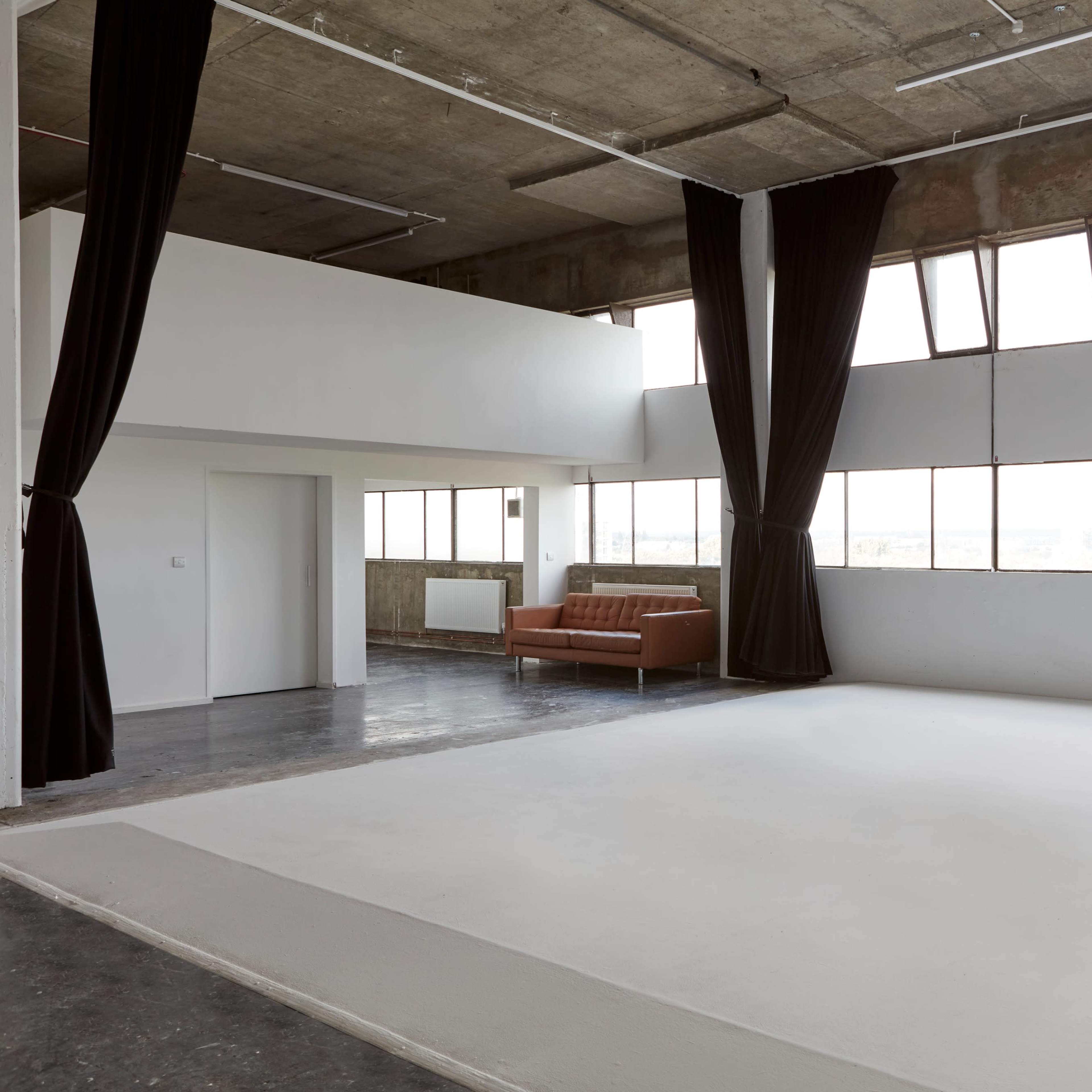 A large, empty studio space features a white floor, high windows, and a brown sofa against one wall.