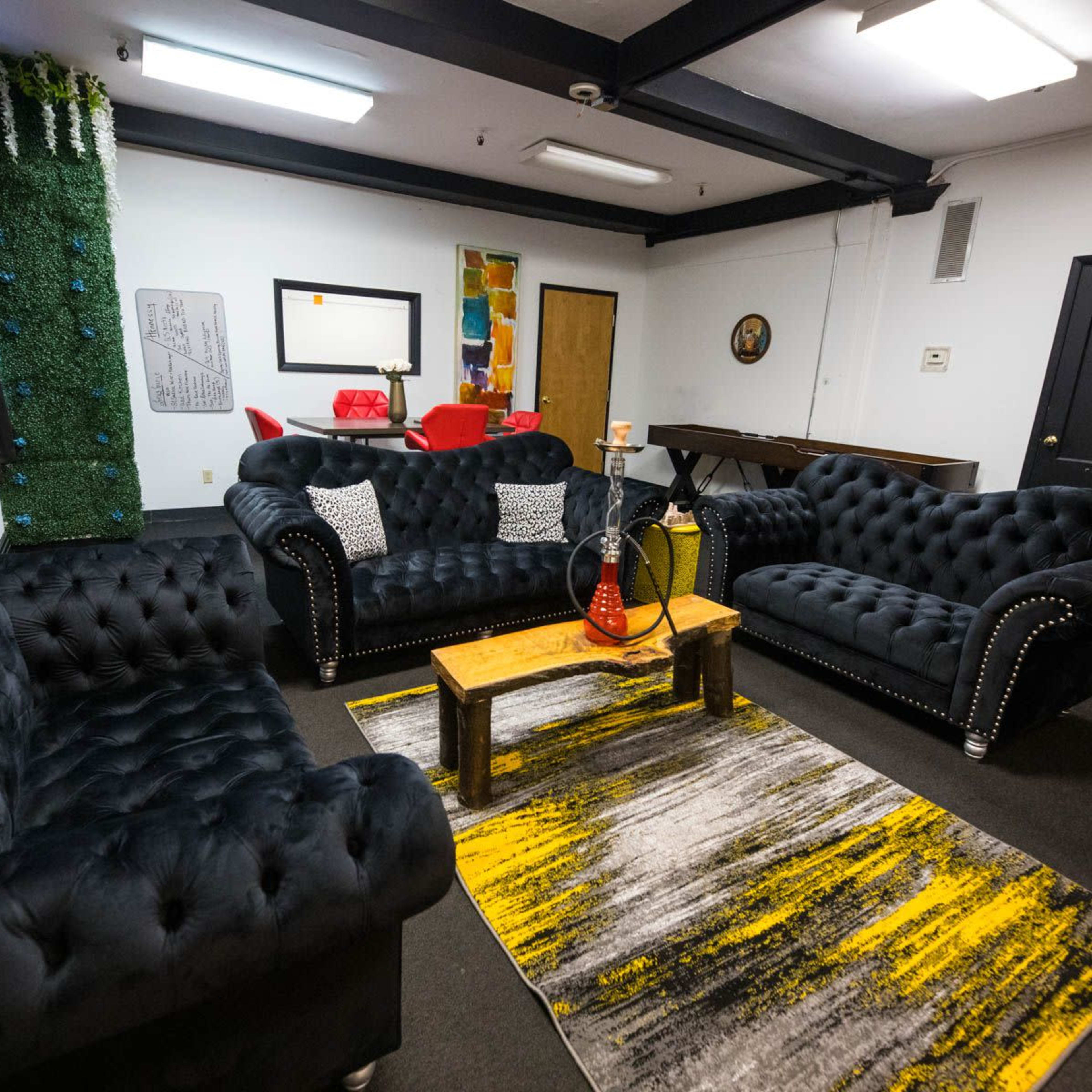 The room features black tufted sofas arranged around a wooden coffee table on a patterned rug, with a mirror and colorful decorations on the walls.