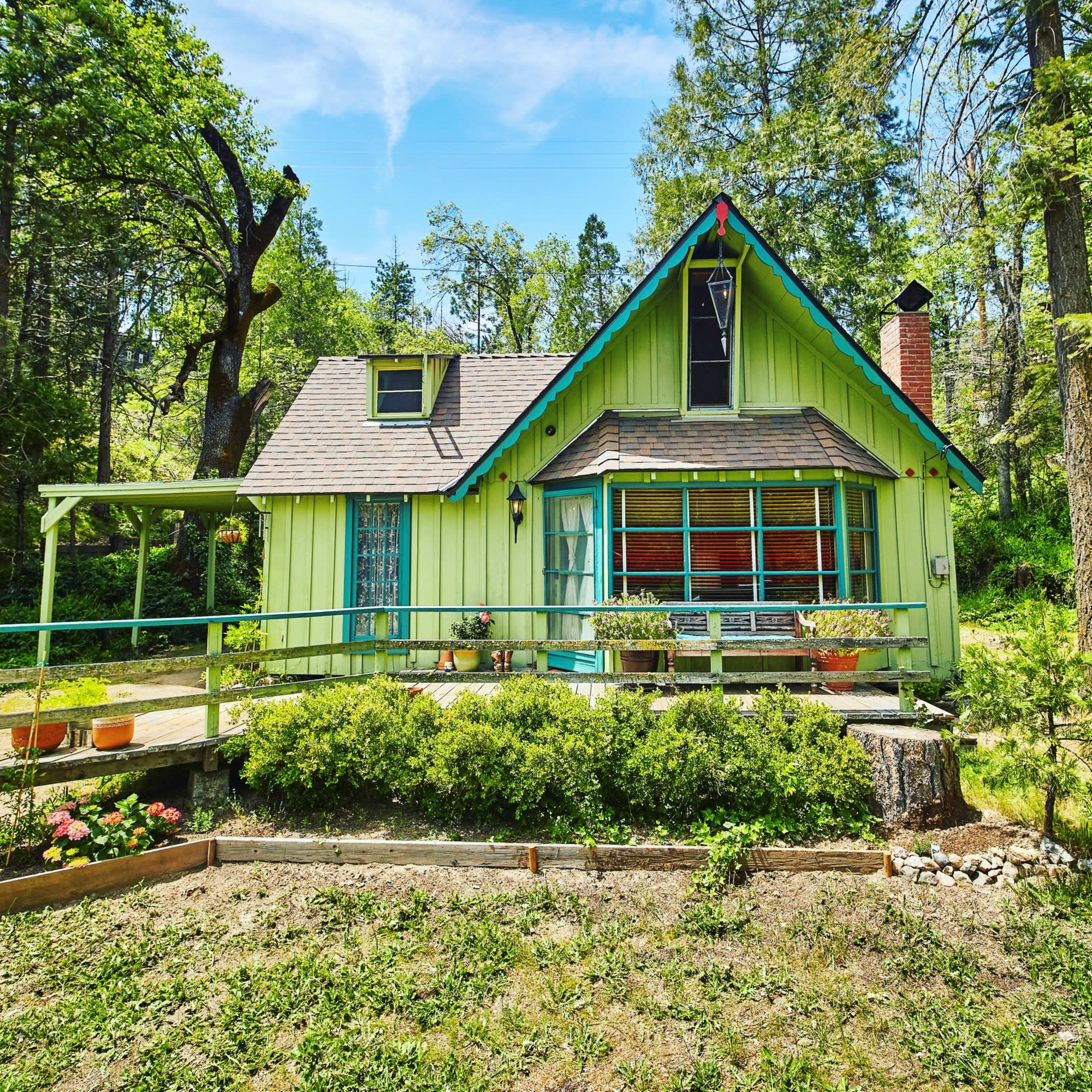 A green, A-frame cottage with large windows sits surrounded by trees and garden beds in a rural setting.