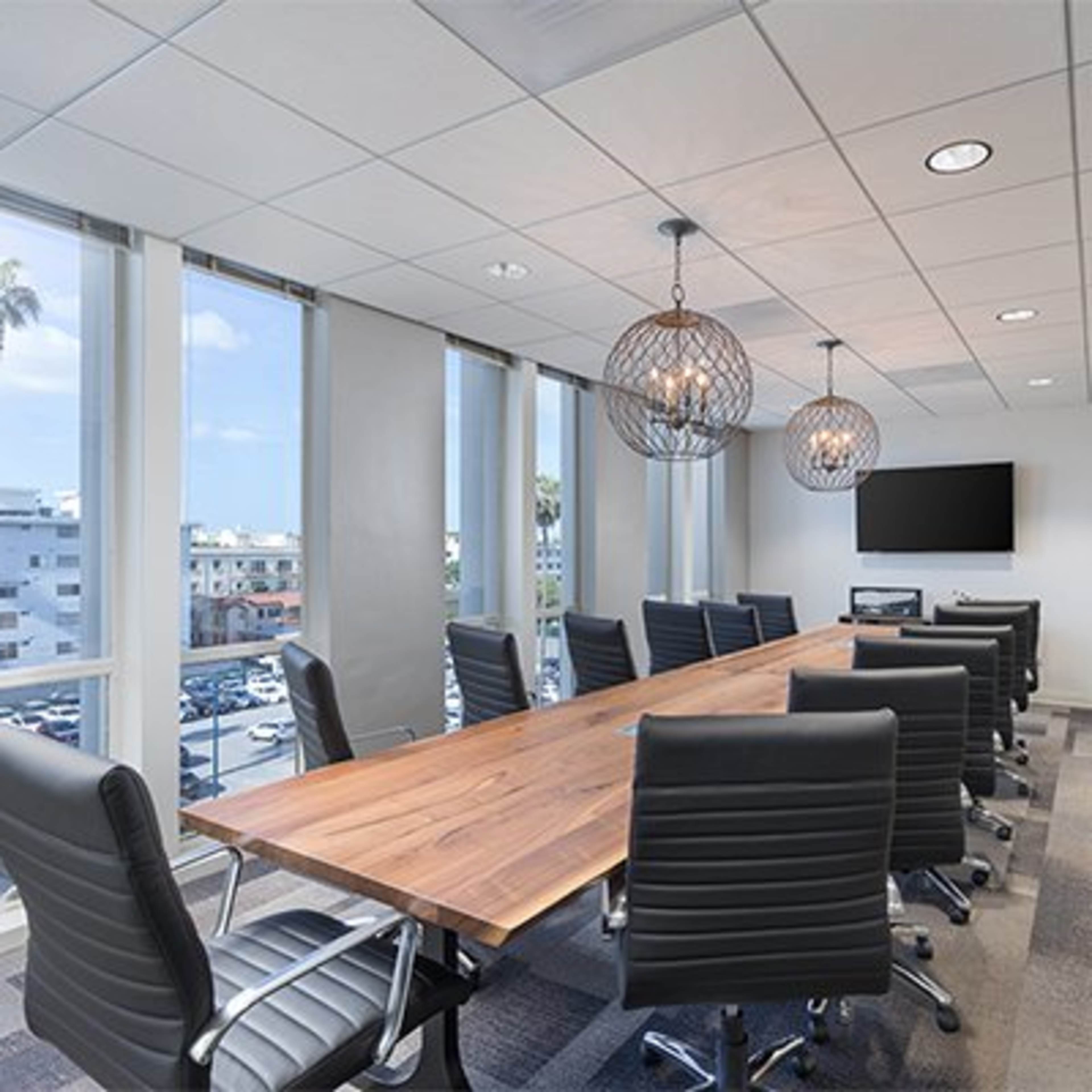 A modern conference room features a large wooden table surrounded by black chairs, with windows offering a view of palm trees and a parking lot outside.