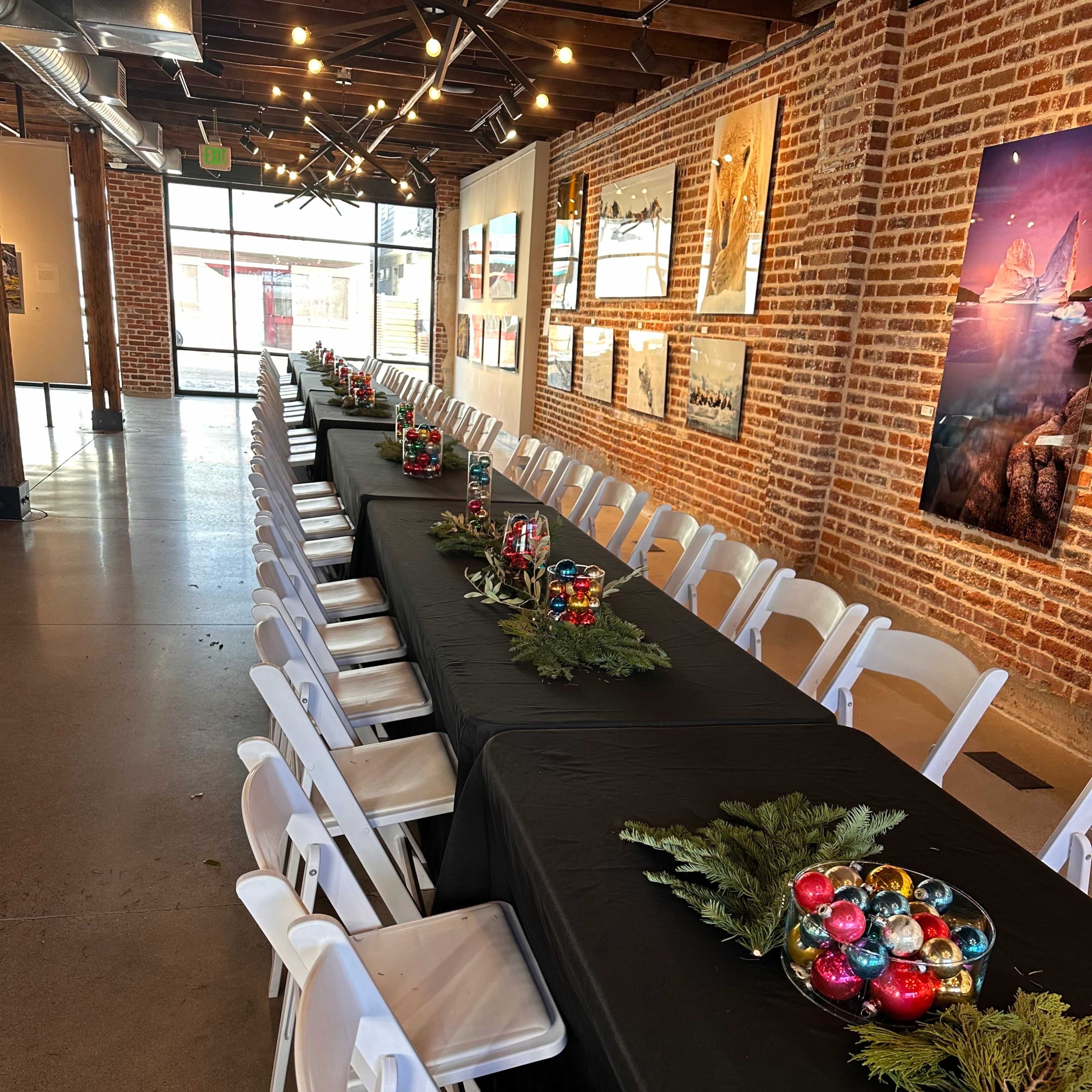 A long table is set with black tablecloths and decorated with colorful ornaments and greenery in a brick-walled event space.