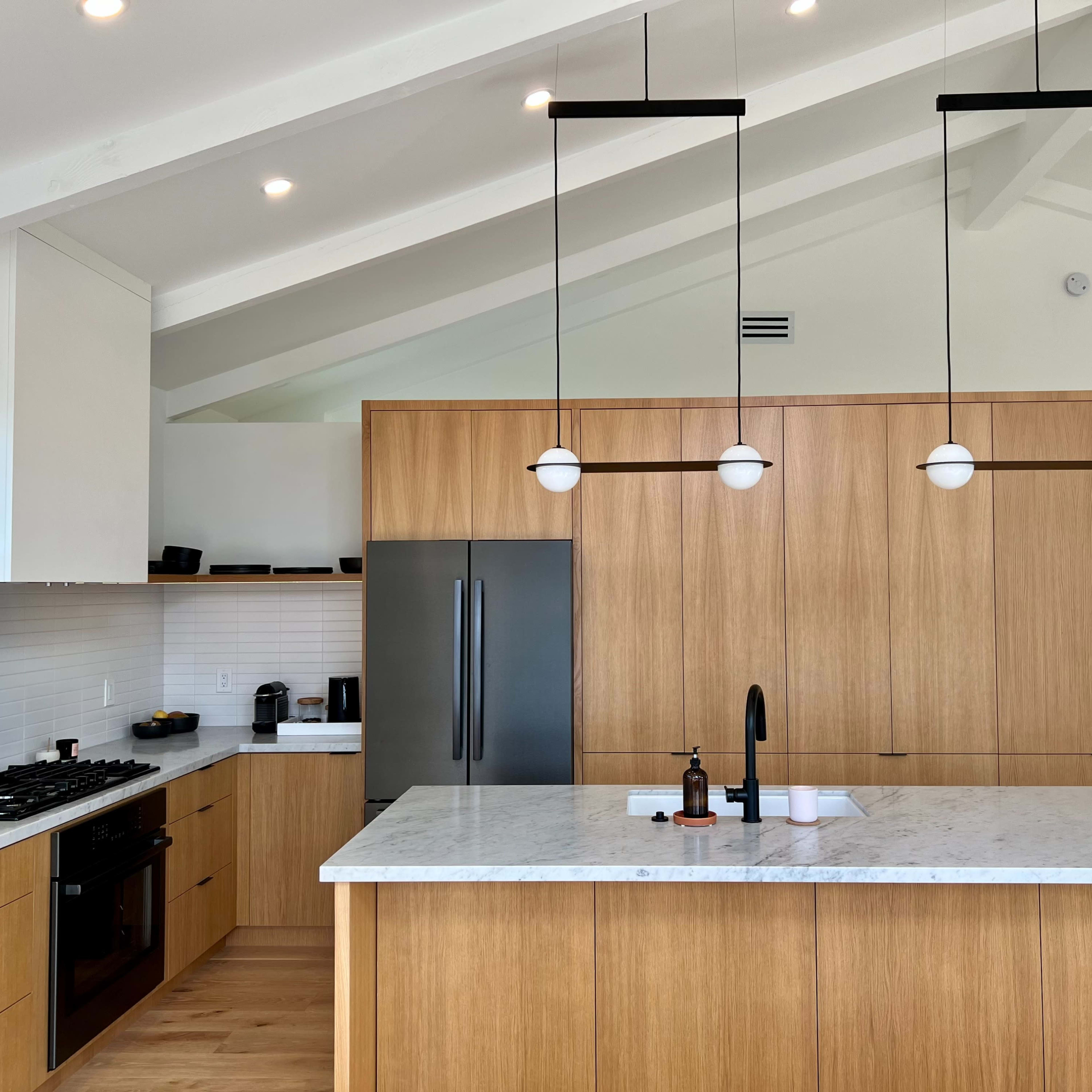A modern kitchen features an island with a marble countertop, wooden cabinetry, and pendant lights hanging from a vaulted ceiling.