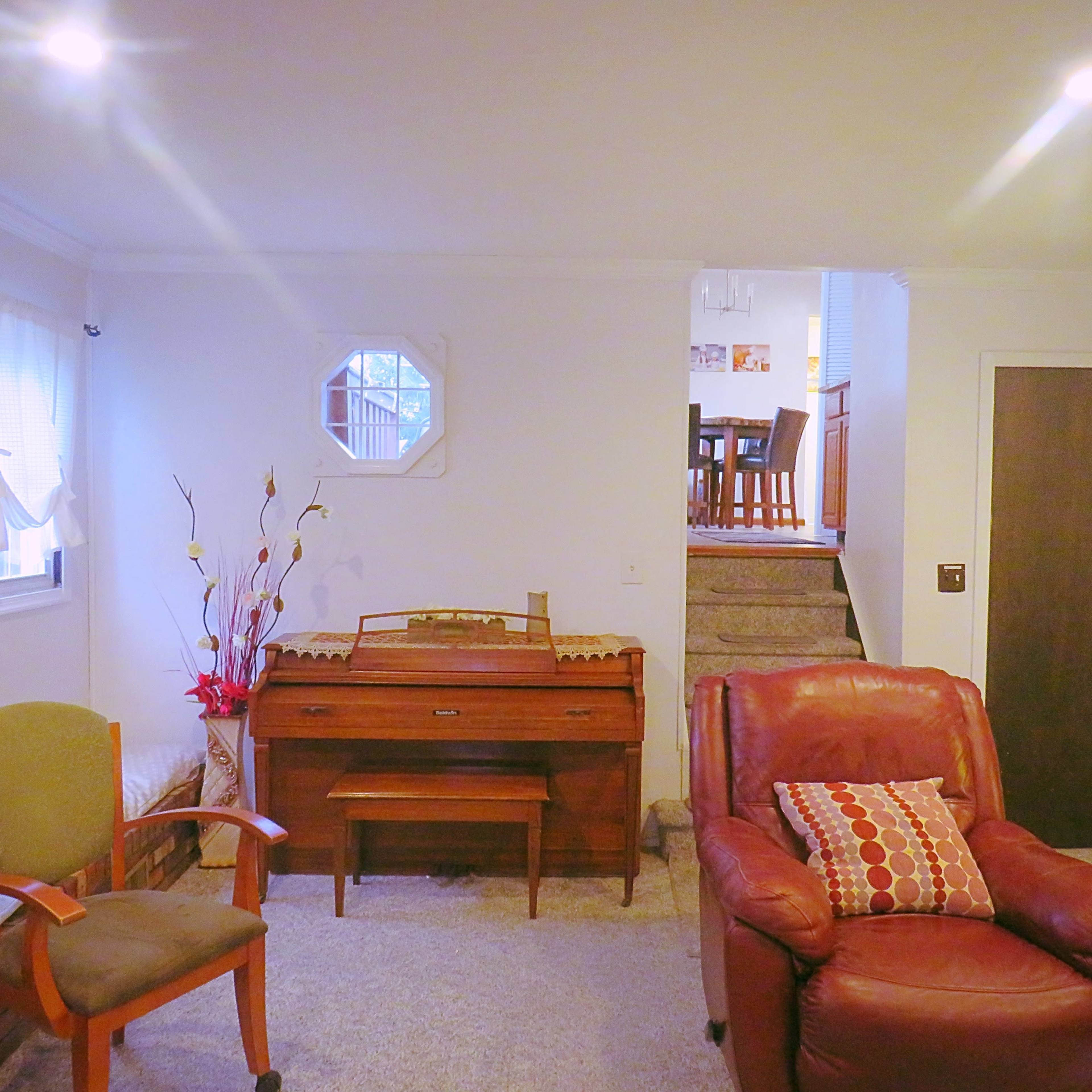 A cozy living room features a wooden piano, a green chair, and a red leather armchair, with stairs visible at the back leading to another floor.