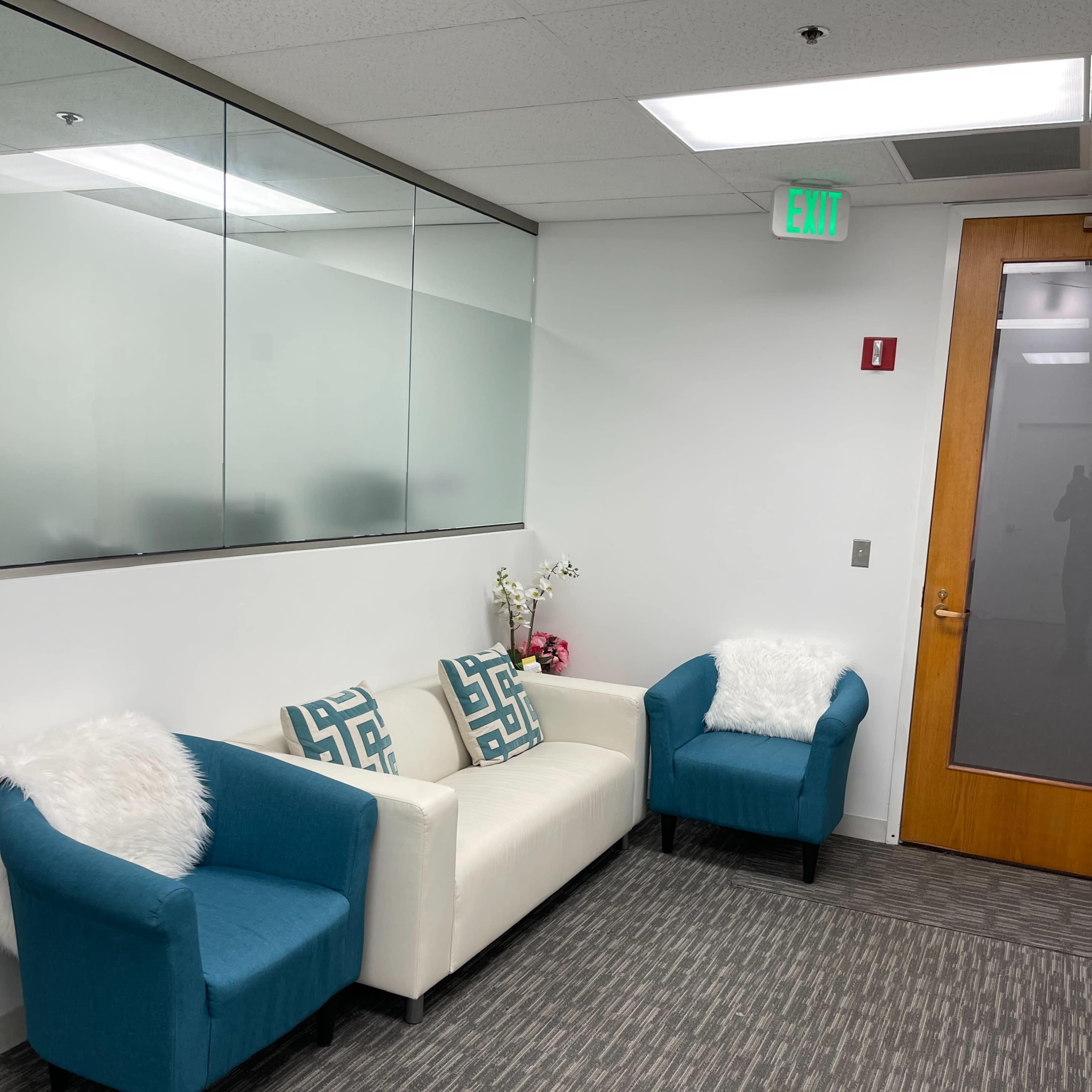 A modern waiting area with a white sofa and two turquoise chairs, adorned with decorative pillows and a small plant on a table.
