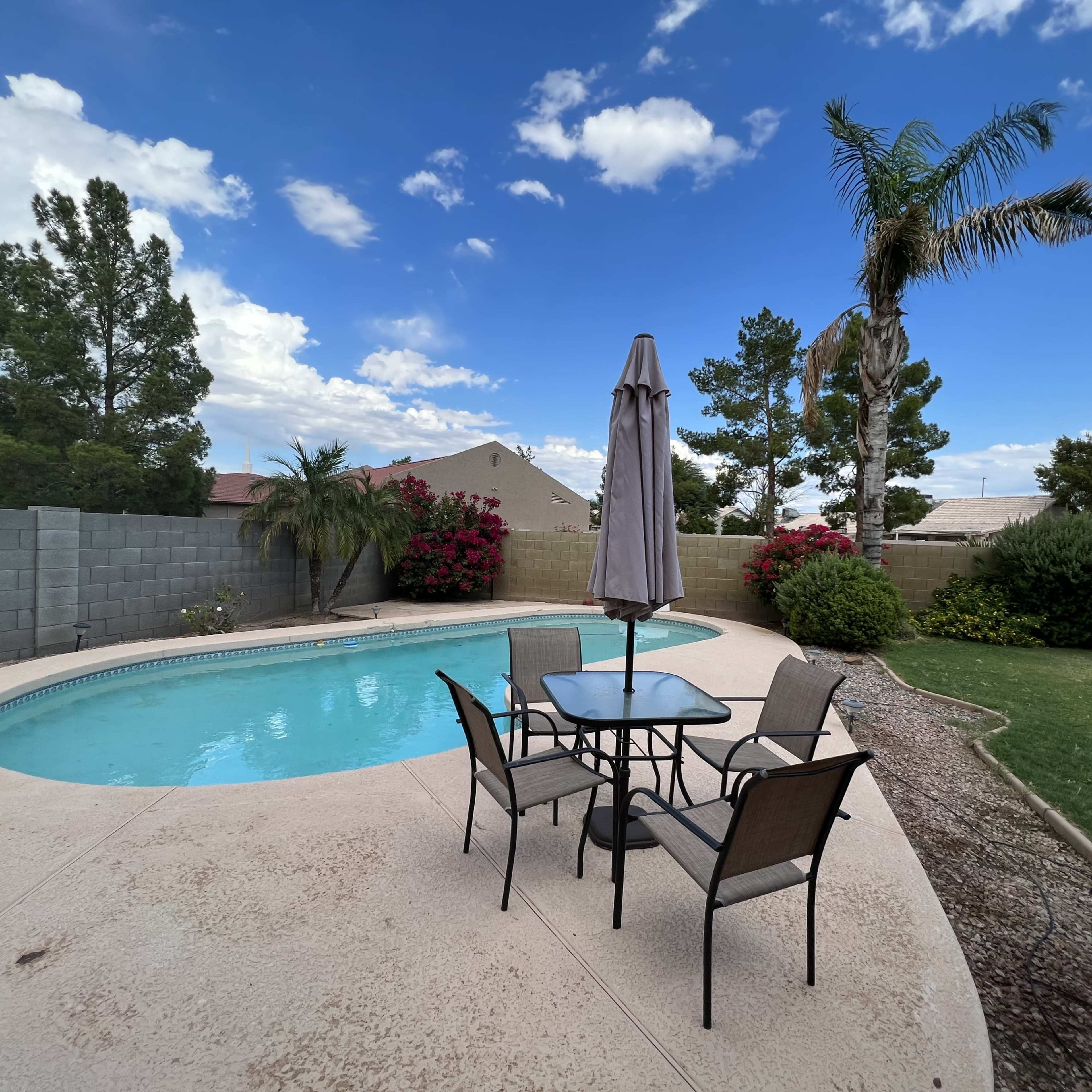 A patio area with a table and chairs is situated beside a swimming pool, surrounded by greenery and flowering bushes under a partly cloudy sky.