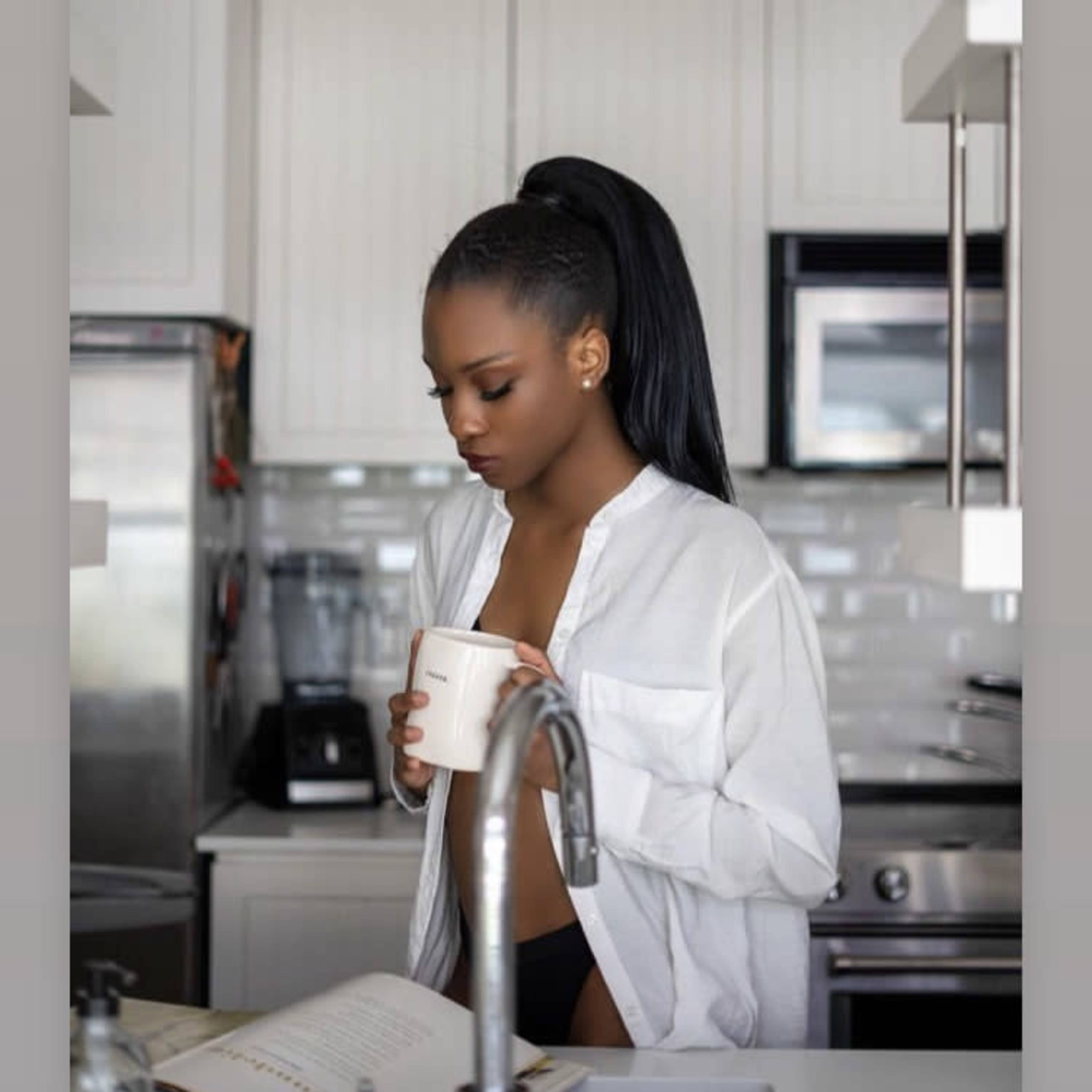 A person stands in a kitchen, holding a cup while looking down at an open book on the counter.