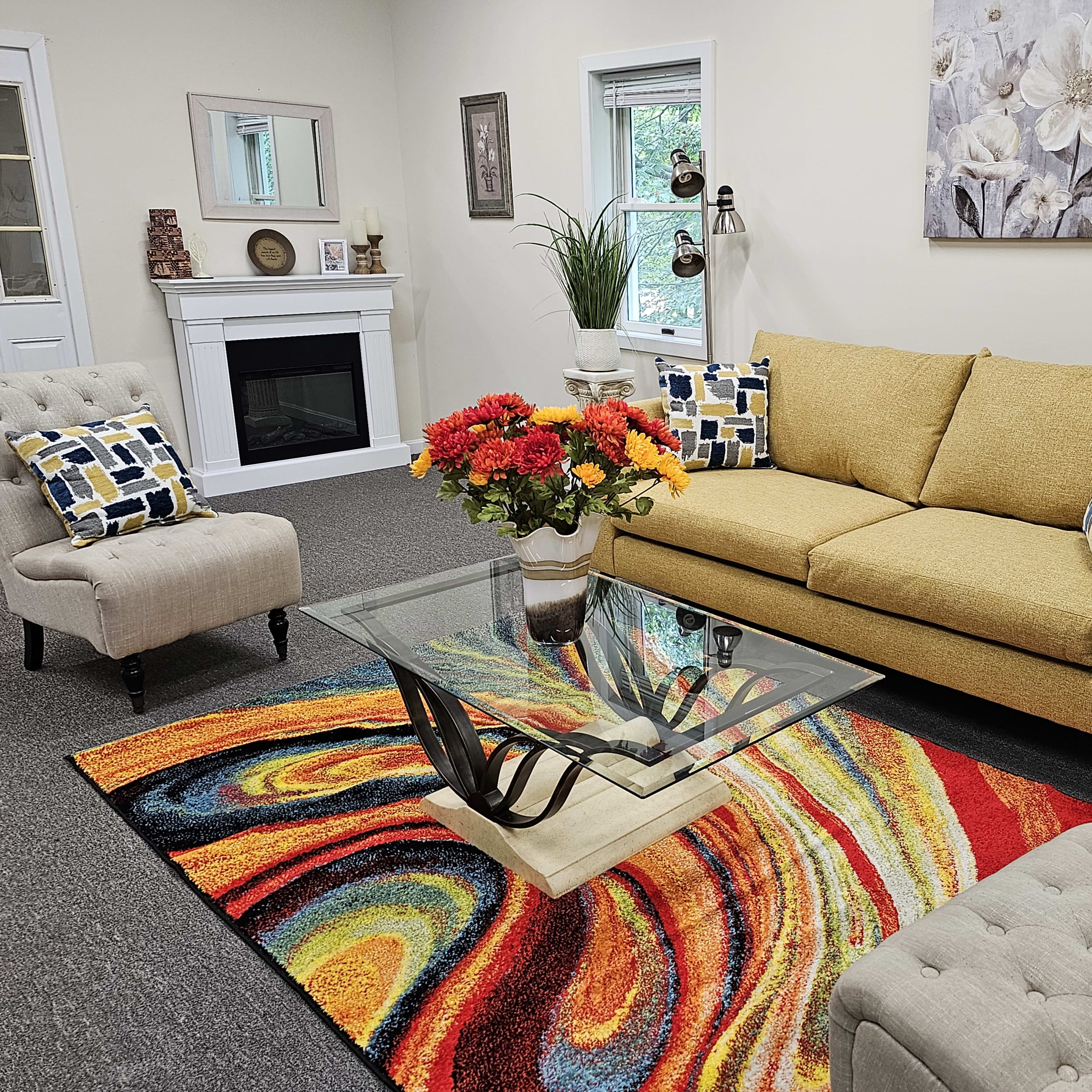 A cozy living room with a yellow sofa, two accent chairs, a glass coffee table, and a colorful swirling rug.