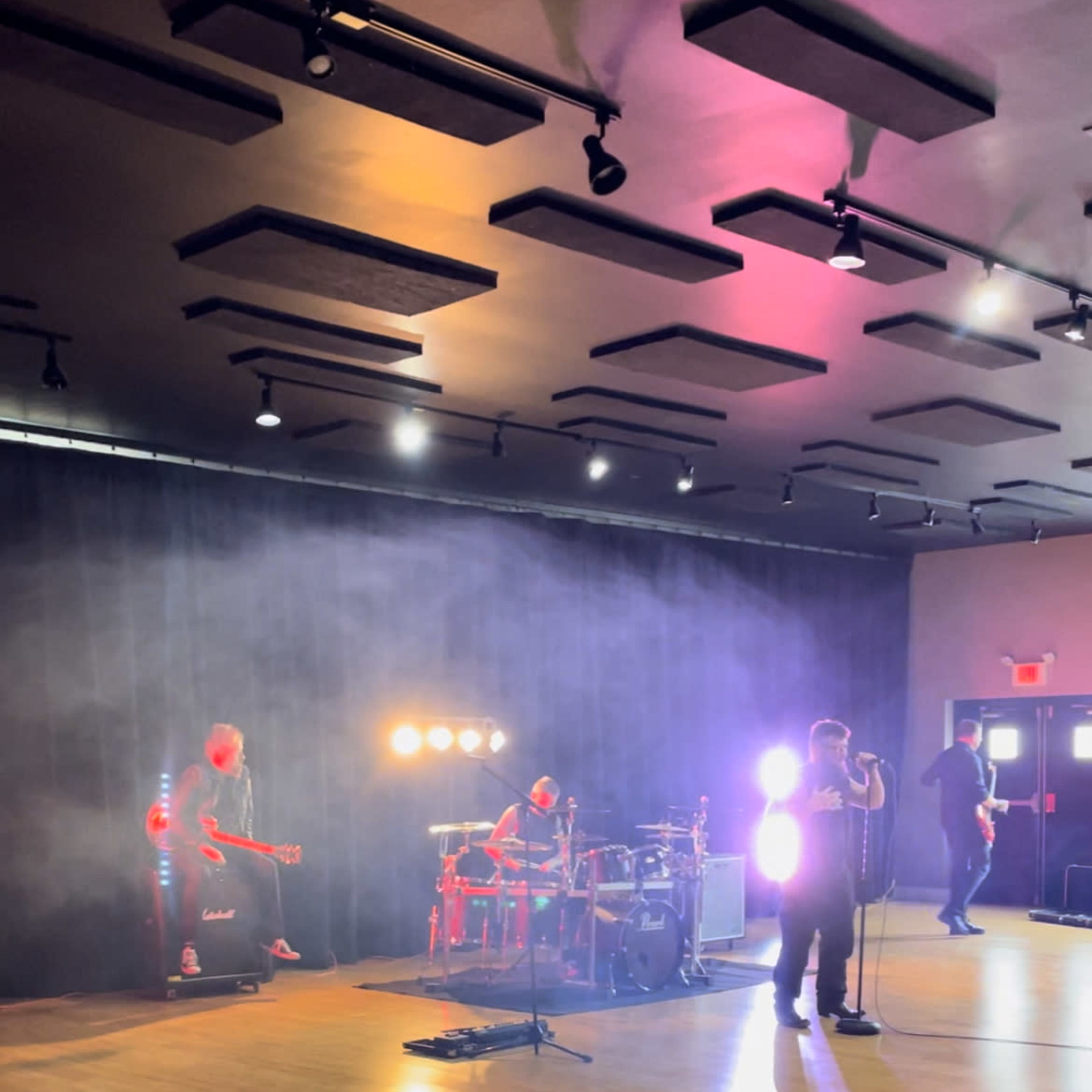 A live band performs on stage with dramatic lighting and a smoke effect in a spacious venue.
