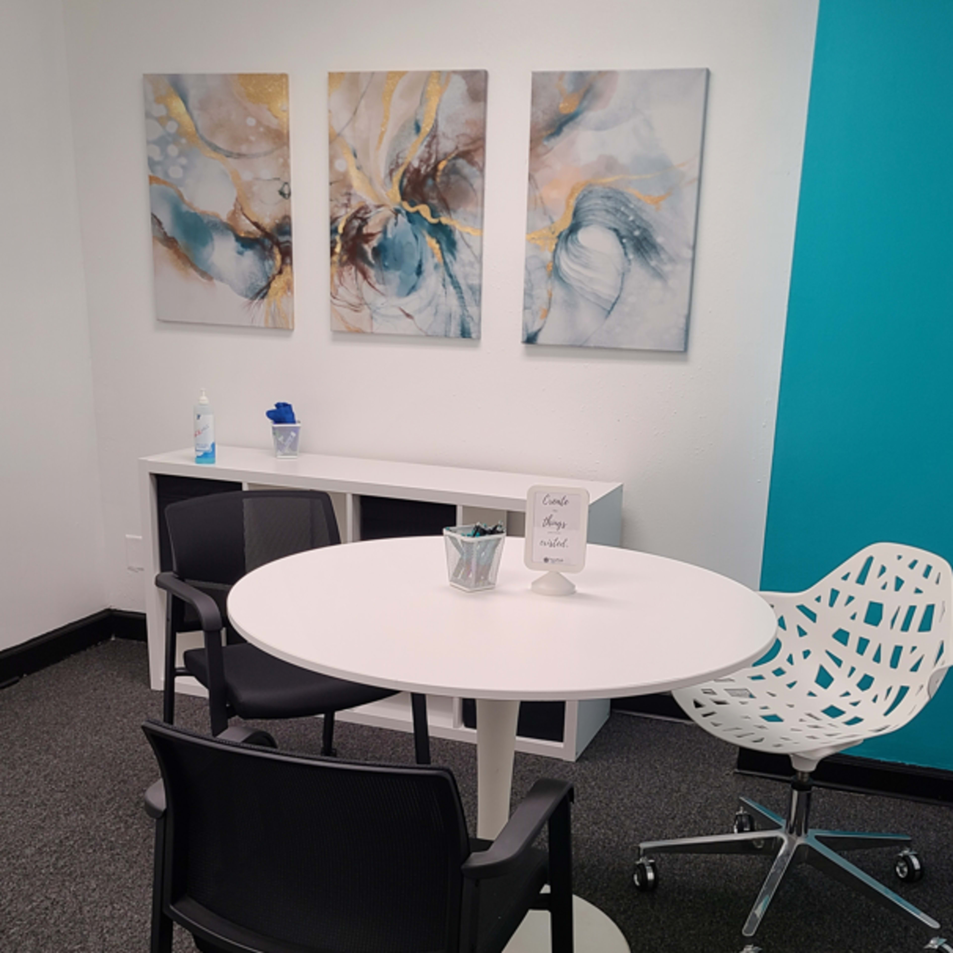A small office space with a round white table, two black chairs, a white decorative chair, a shelf with a blue container, and three abstract paintings on the wall.