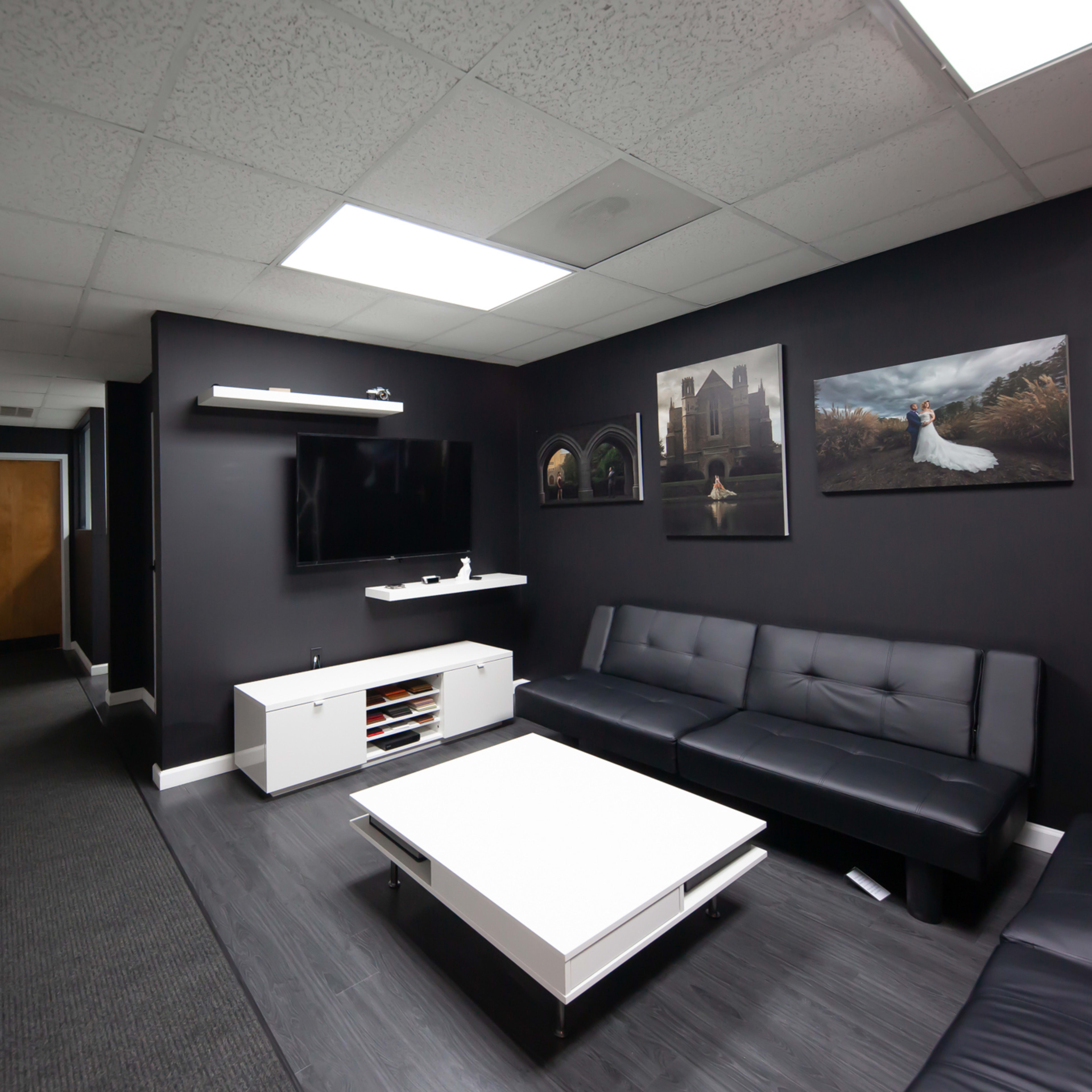 A modern office space with dark walls, a black sectional sofa, a white coffee table, and a television mounted on the wall, along with framed pictures displayed.