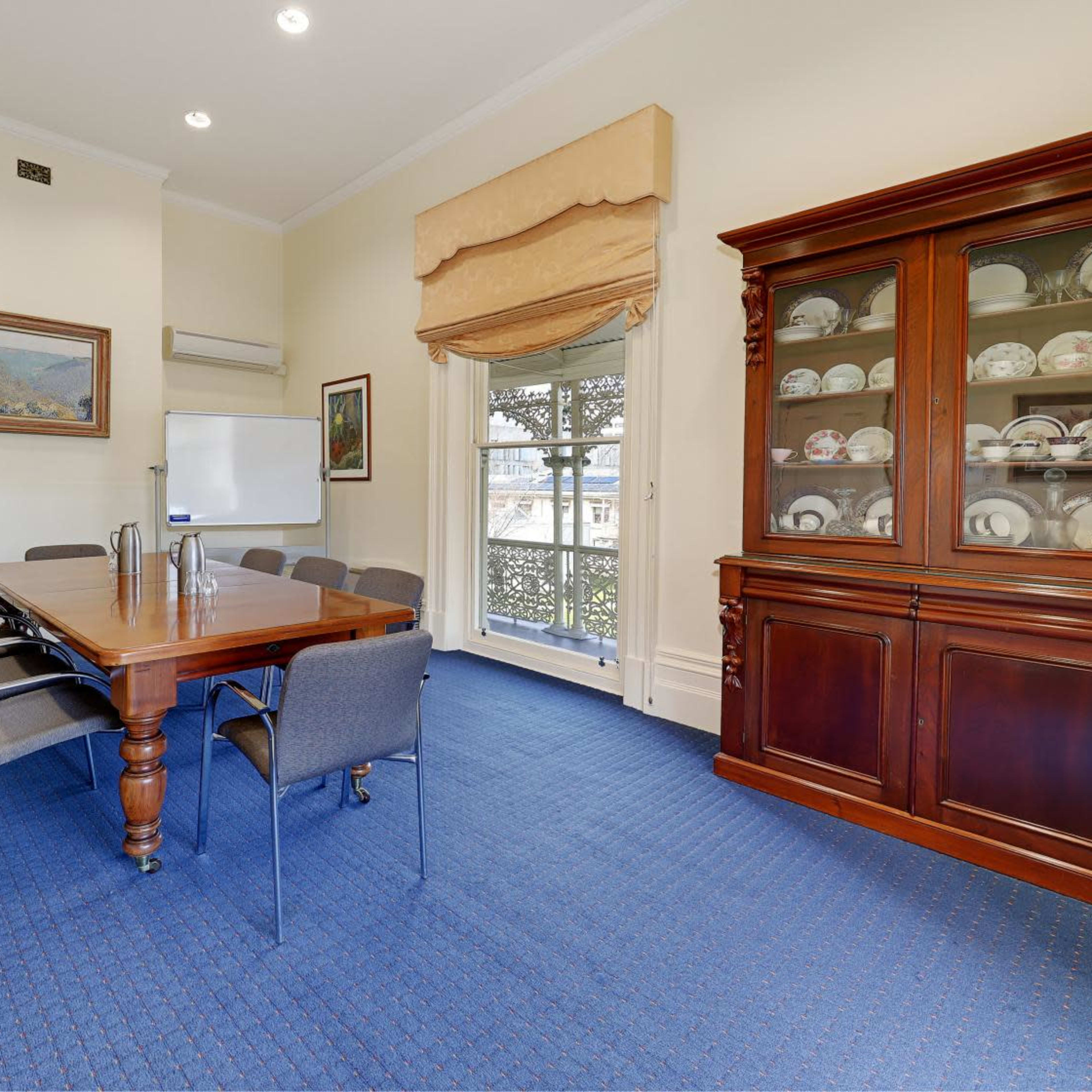 A conference room with a large wooden table, six gray chairs, and a cabinet displaying porcelain plates, illuminated by natural light from a window.