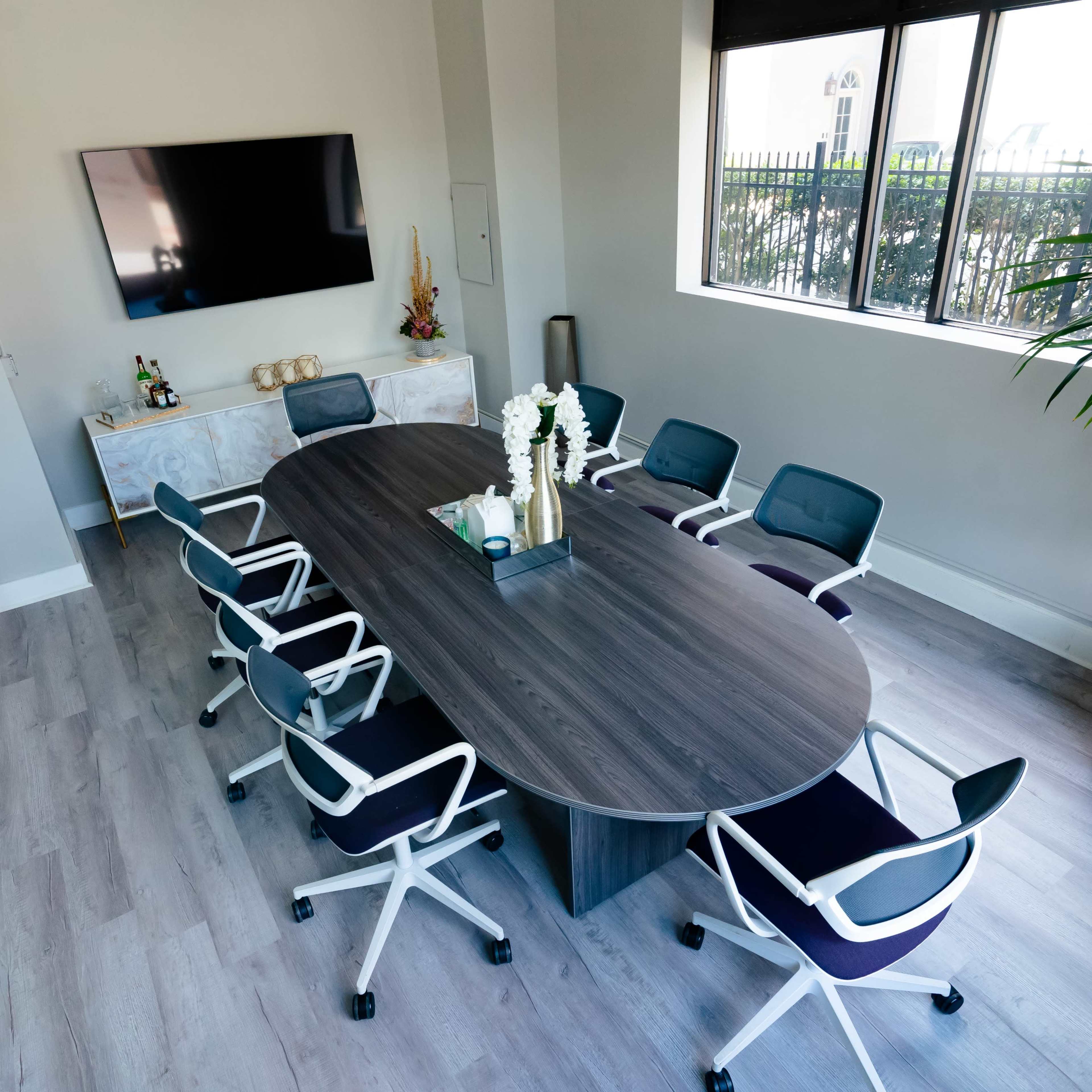 A modern conference room features an oval table surrounded by eight office chairs, with a large screen mounted on the wall and a decorative plant in the corner.