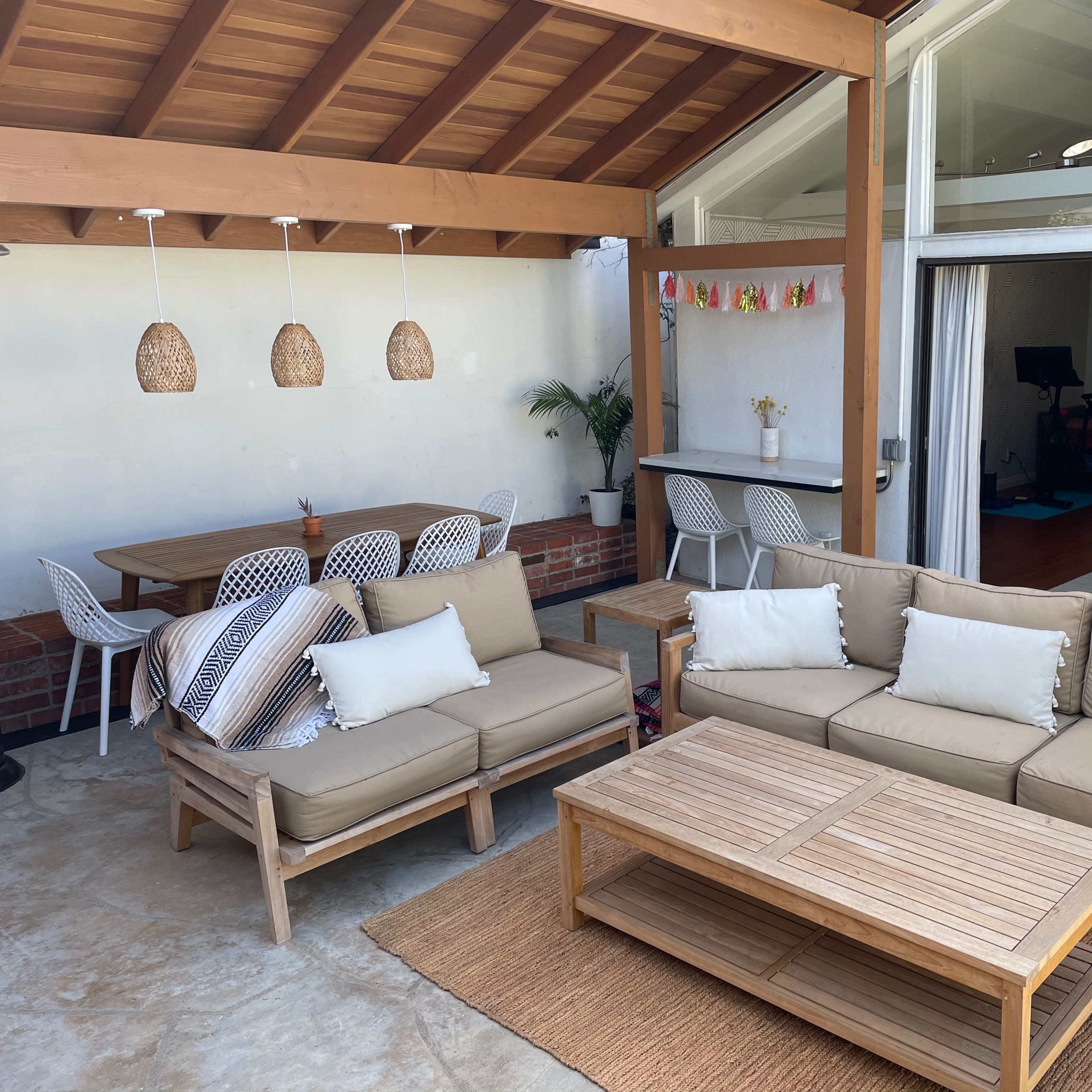 A well-furnished outdoor living space with a beige sectional sofa, a wooden coffee table, and a dining area with white chairs under a wooden ceiling.