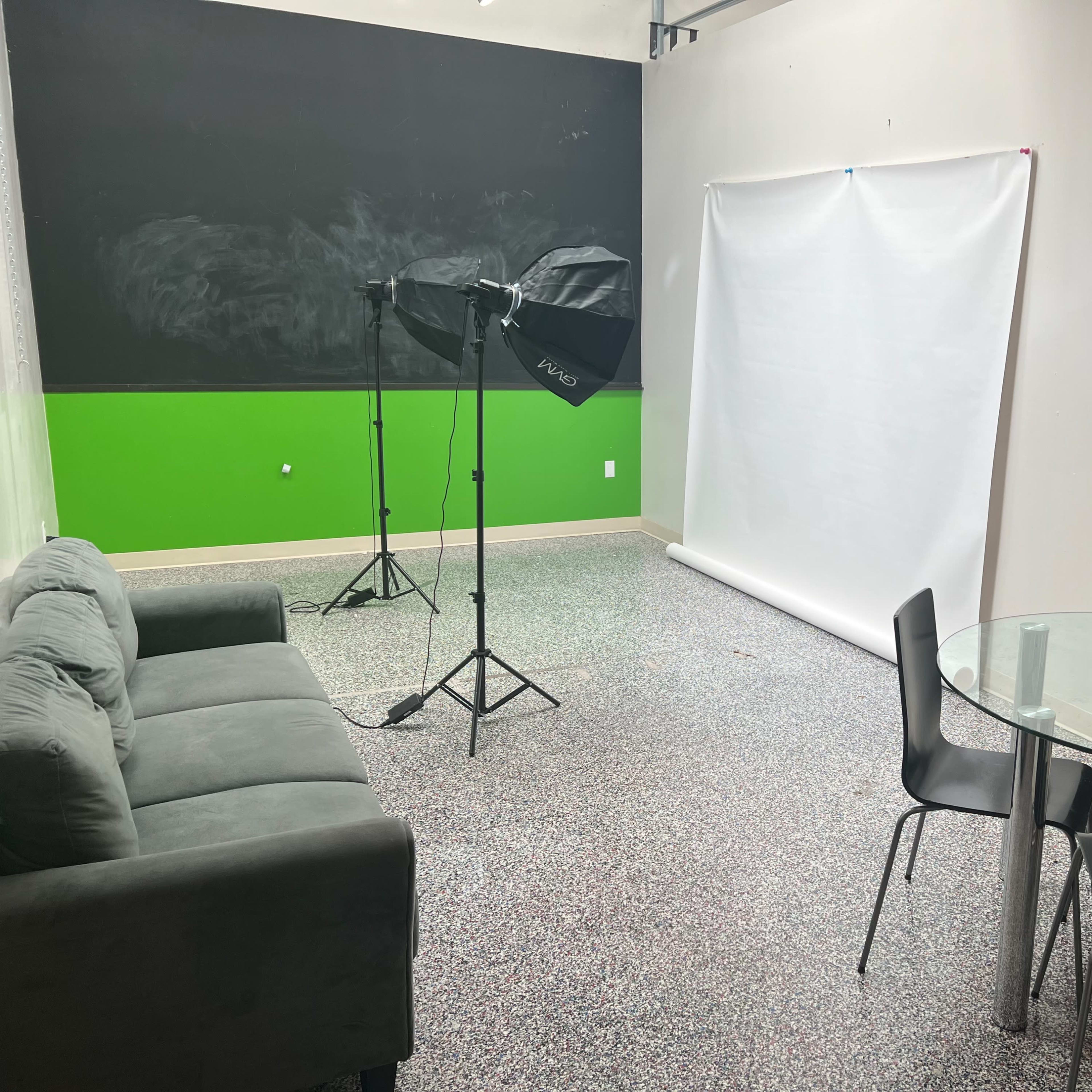A small studio space with a gray couch, a round glass table with four black chairs, and lighting equipment set up near a white backdrop against a green wall.