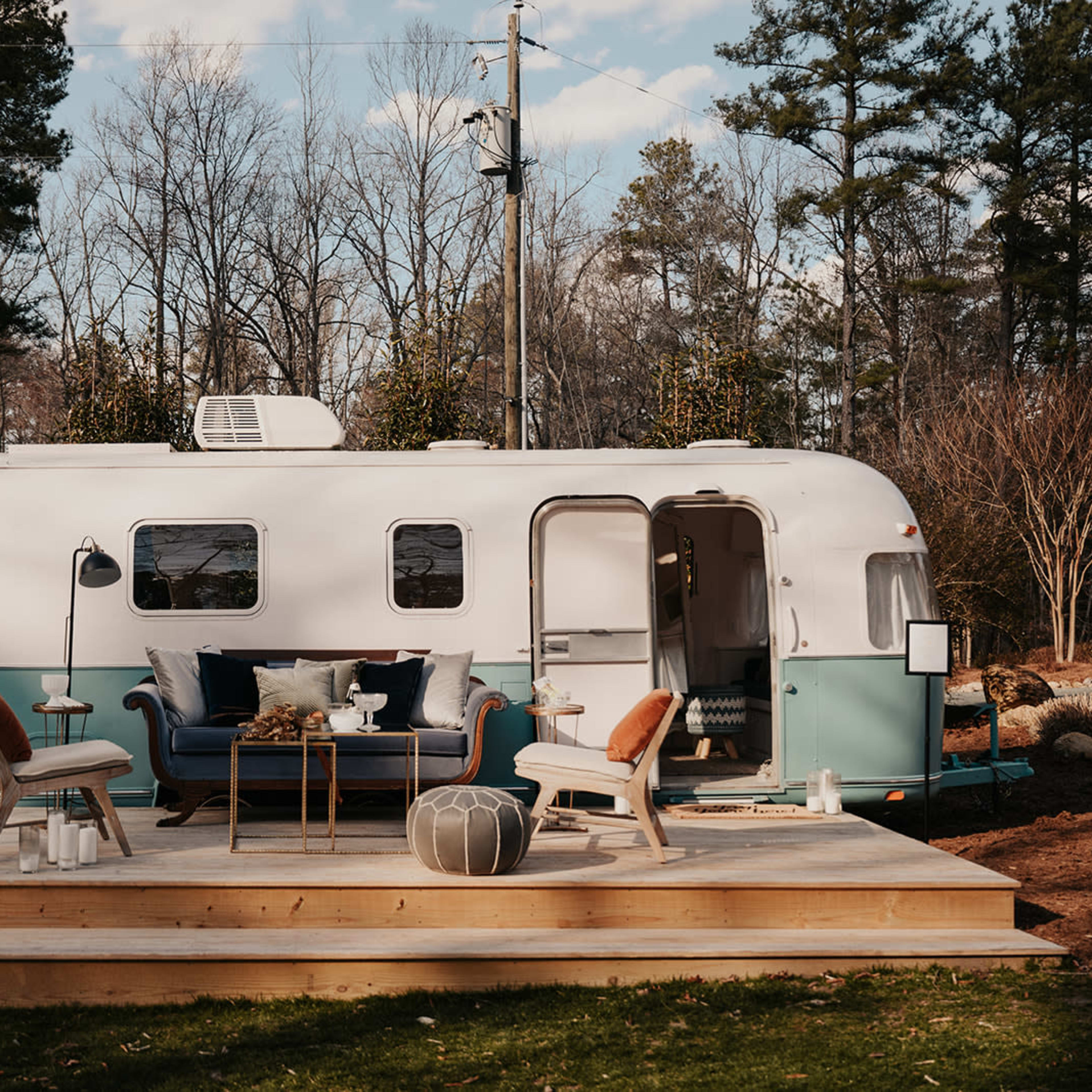 A vintage trailer is parked on a wooden deck in a wooded area, surrounded by trees.