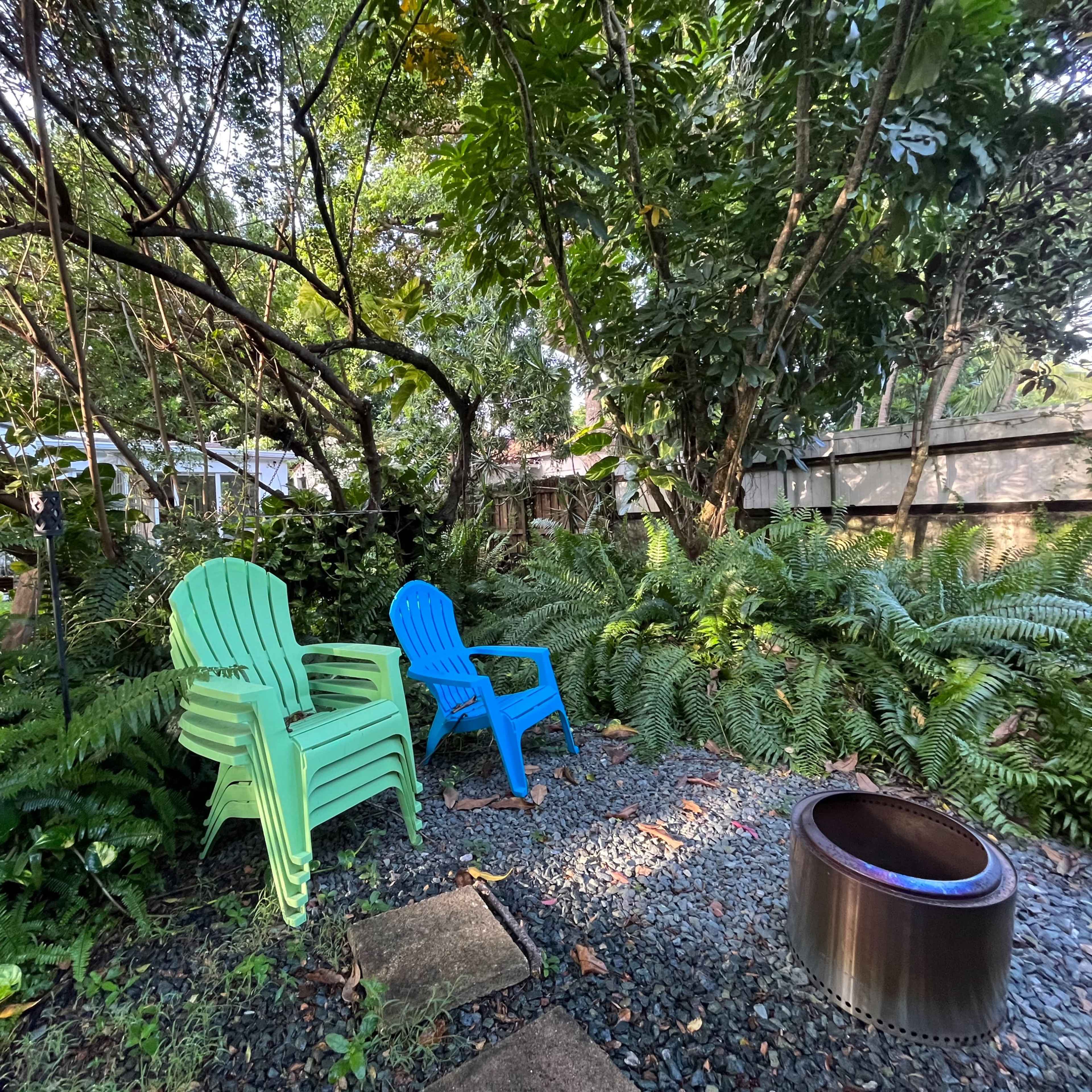 Two colorful adirondack chairs are positioned among lush greenery near a circular fire pit.