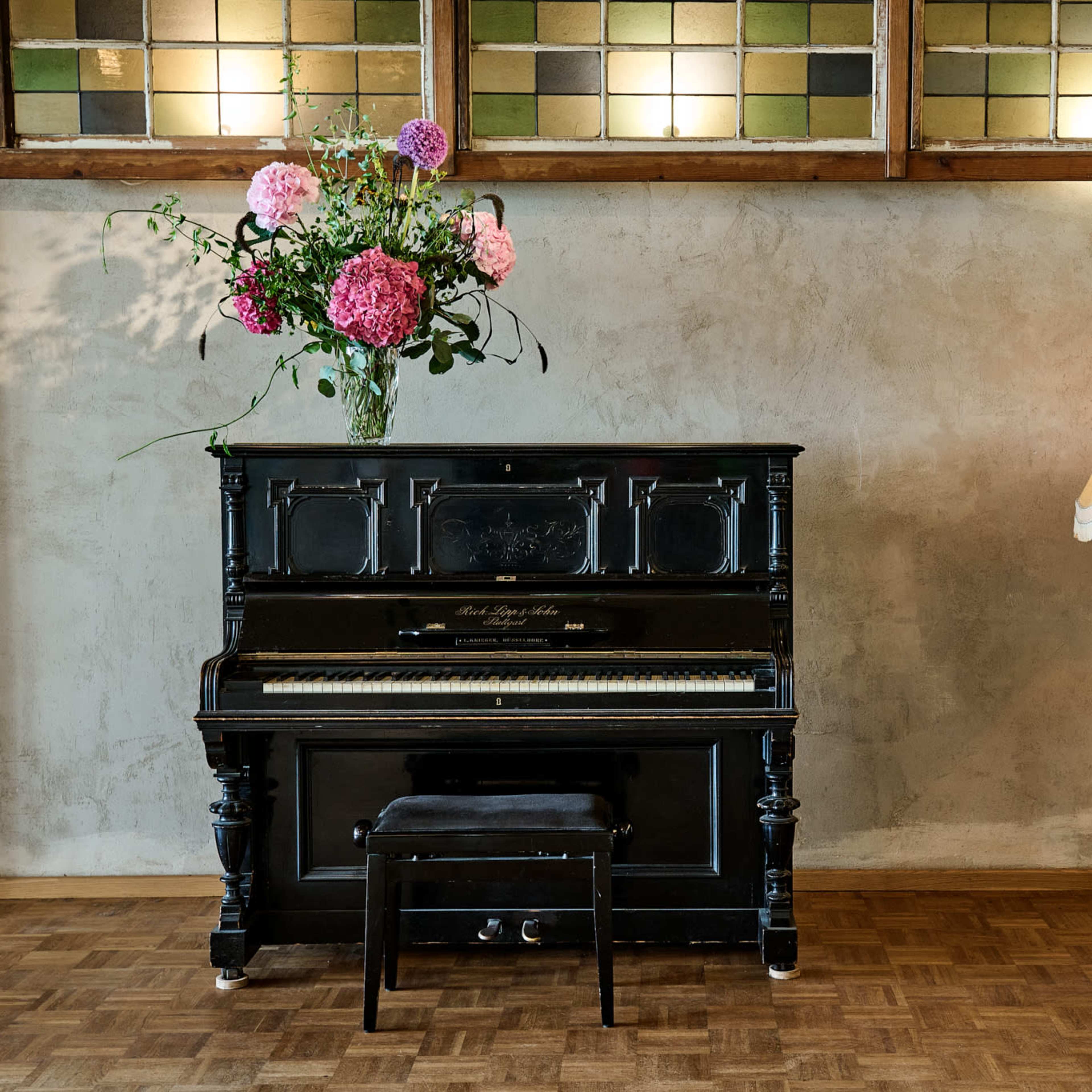 A black piano with a floral arrangement atop it stands next to a gold lamp on a wooden floor against a textured wall.