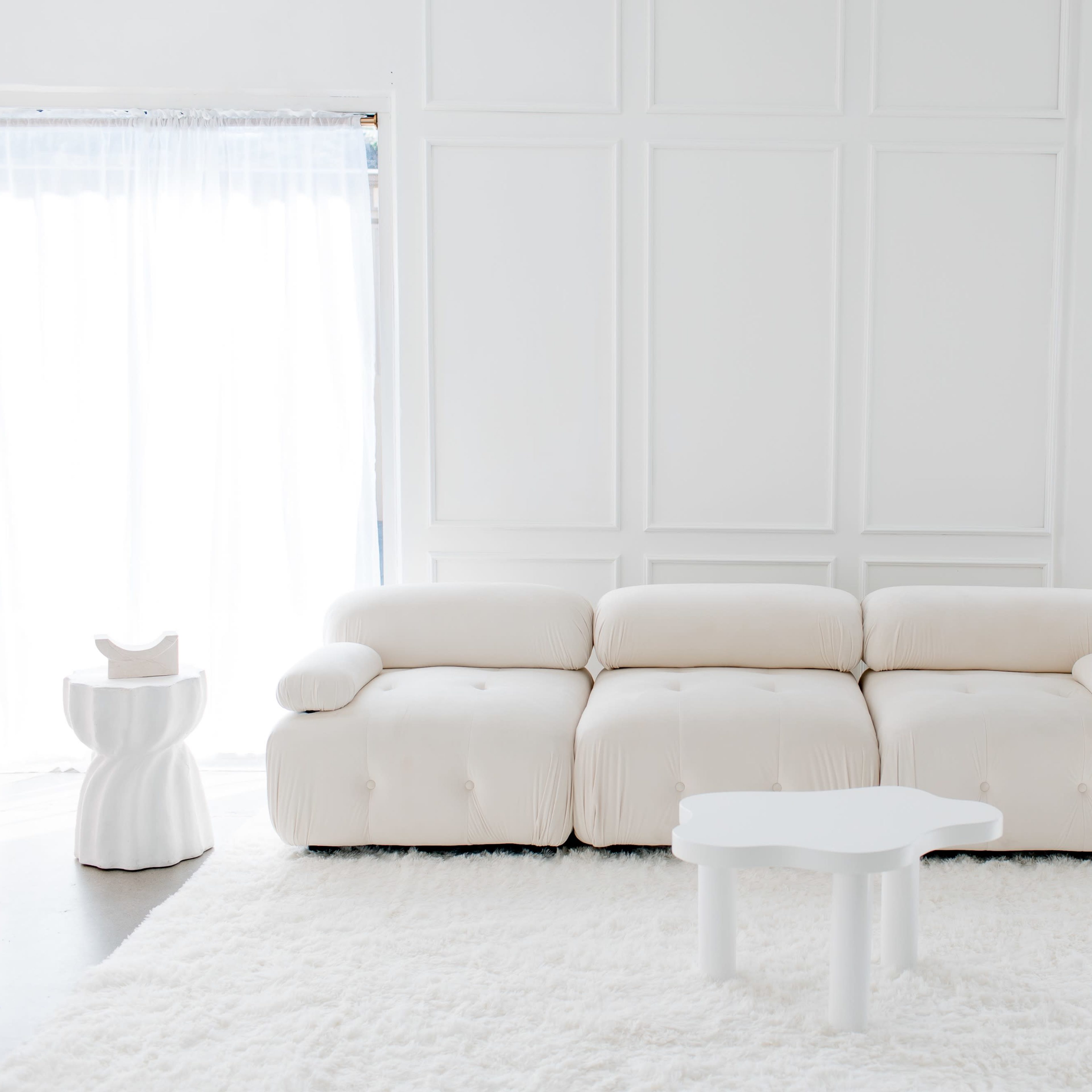 A light-colored, contemporary sofa set in a bright room with a white rug, a small side table, and minimalist decor elements.